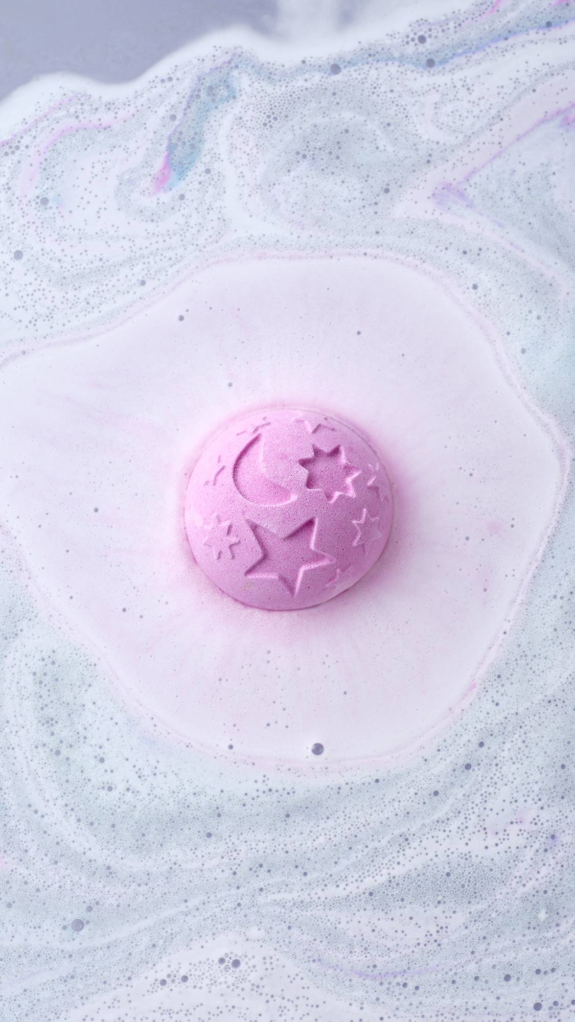 A top-down view shows the Twilight Bath Bomb dissolving, creating a soft pink halo in the water. The lavender sphere, featuring embossed celestial designs, floats amidst a sea of frothy white and light blue foam.