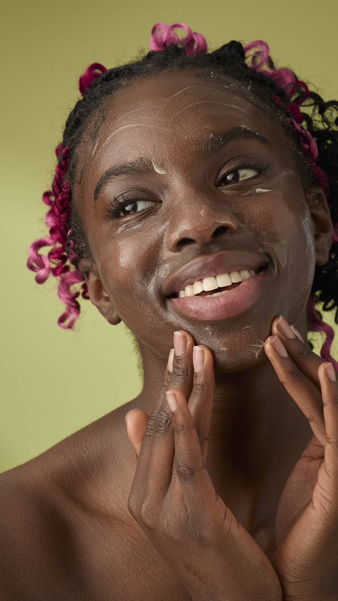 A person with pink braids smiles while gently massaging Ultrabland cleanser onto their jawline and cheeks against an olive green background.