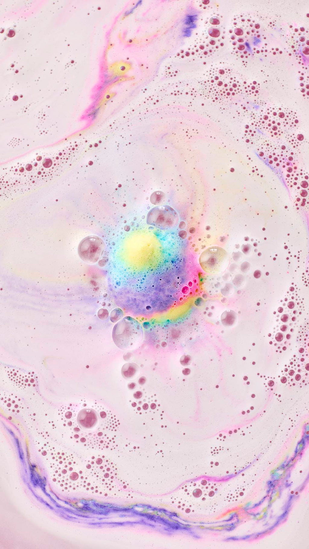 A top-down view shows the colorful Unicorn Poop Bath Bomb dissolving in the center of the tub. It releases a vibrant mix of yellow, blue, and pink foam into milky, pastel purple water, creating a mesmerizing pattern of swirling colors and bubbles.