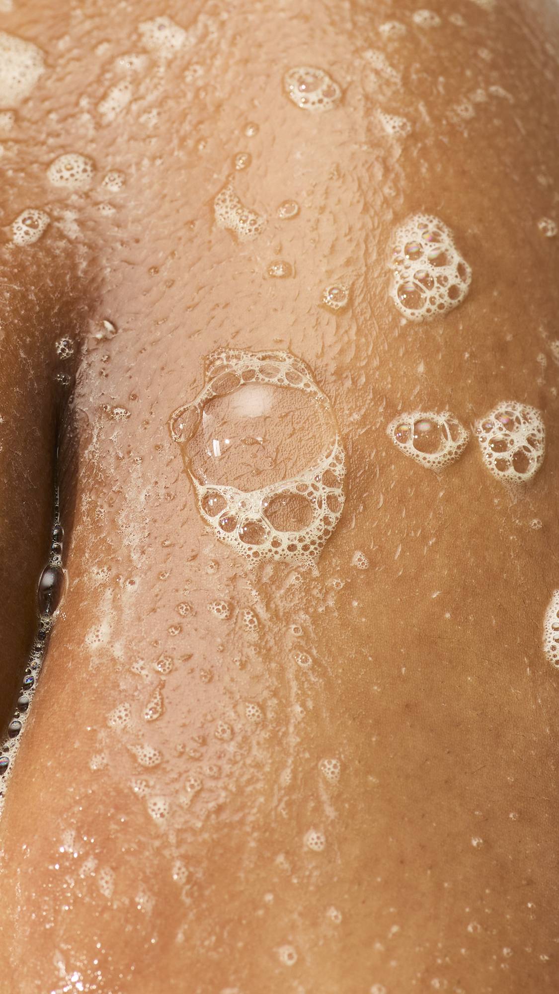 A close-up of wet, glistening skin in the shower, covered in soapy lather and clusters of translucent bubbles.