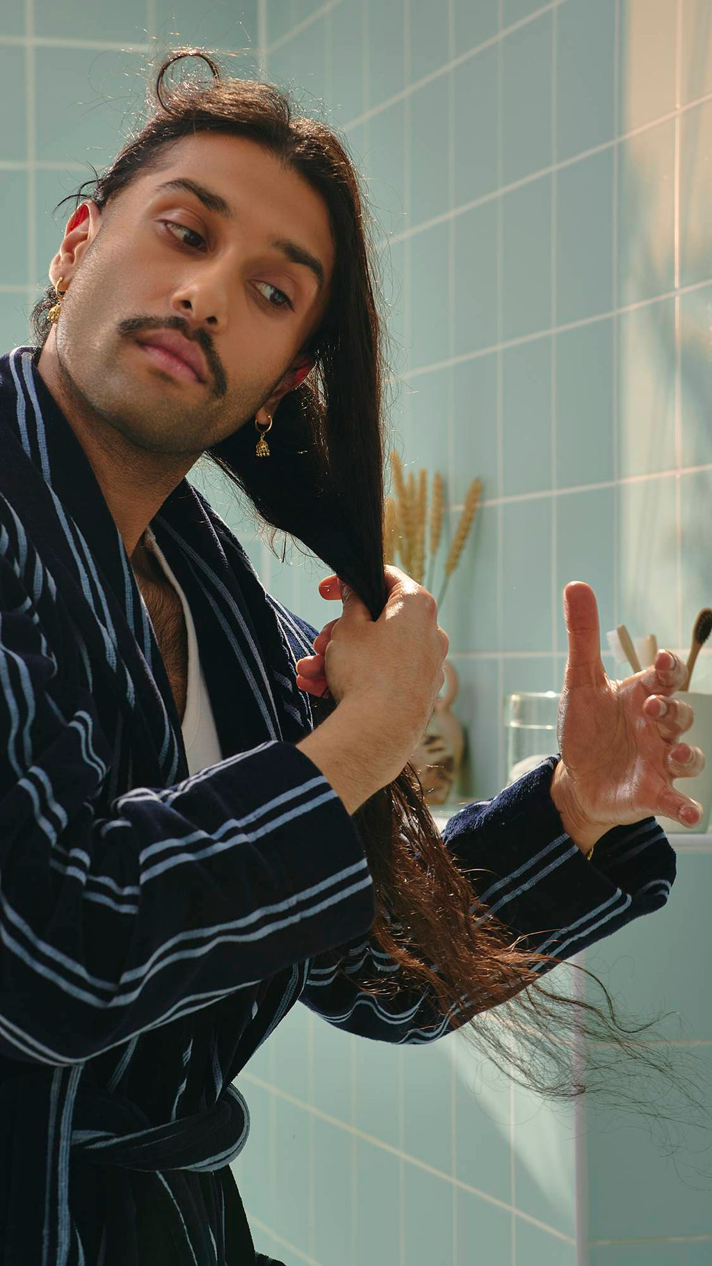 A model with long dark hair and a moustache, wearing a navy and white striped dressing gown, runs their hands through their hair to apply the "Vegan Leather Jacket" hair and beard oil. Light green tiles are visible in the background.