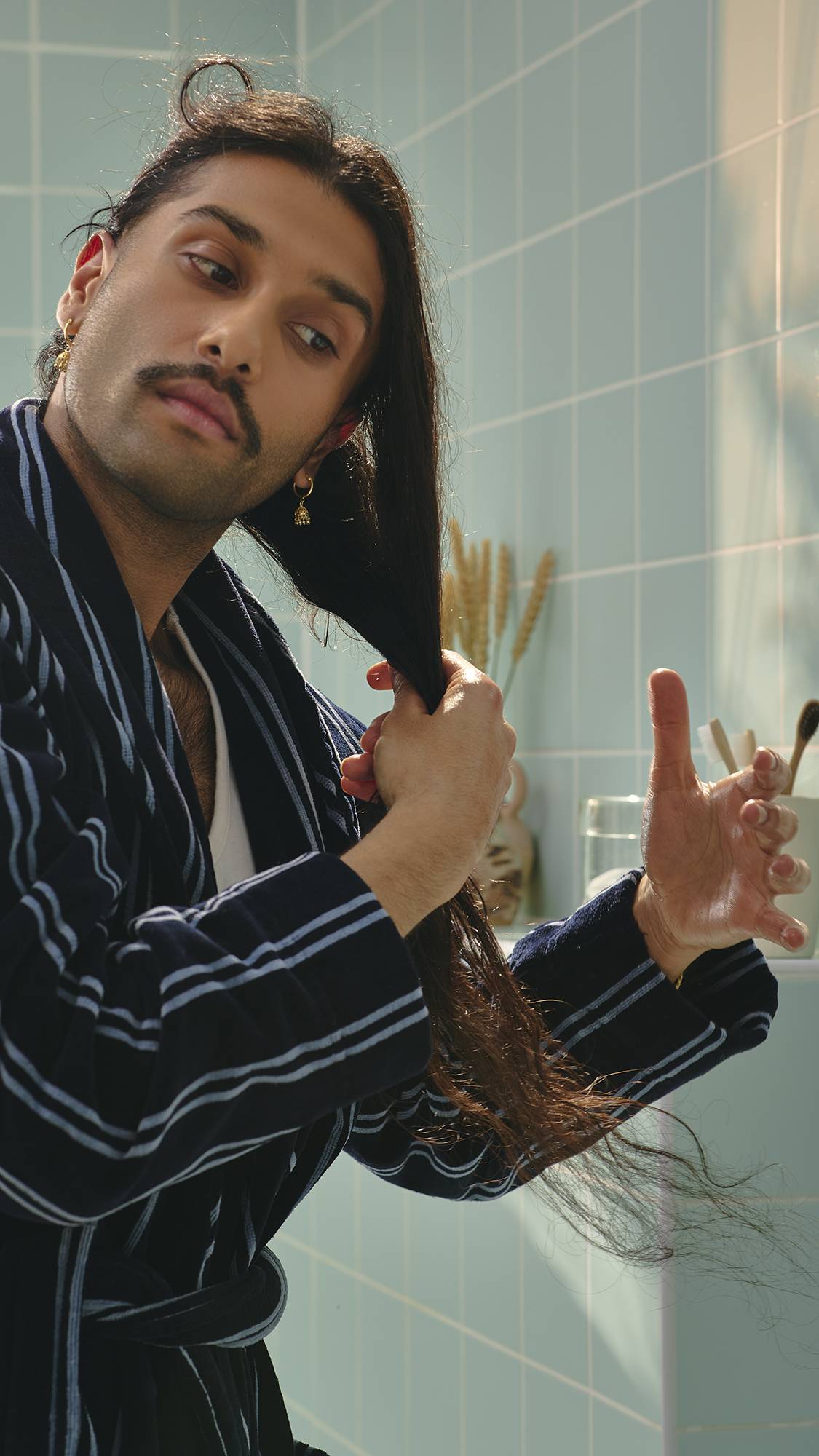 A model with long dark hair and a moustache, wearing a navy and white striped dressing gown, runs their hands through their hair to apply the "Vegan Leather Jacket" hair and beard oil. Light green tiles are visible in the background.