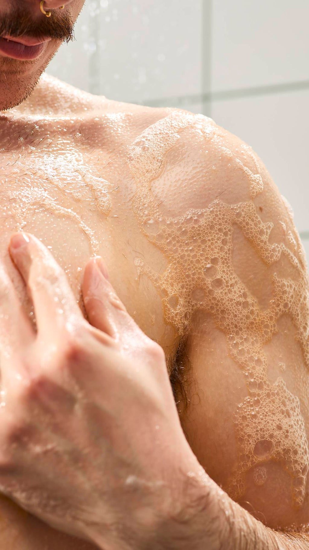 A close-up of a person's chest and shoulder covered in a warm orange soapy lather with small bubbles in the shower. A hand rests on the wet skin.
