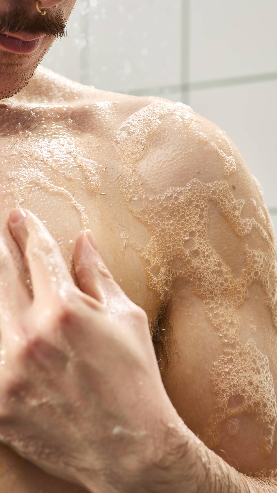 A close-up of a person's chest and shoulder covered in a warm orange soapy lather with small bubbles in the shower. A hand rests on the wet skin.
