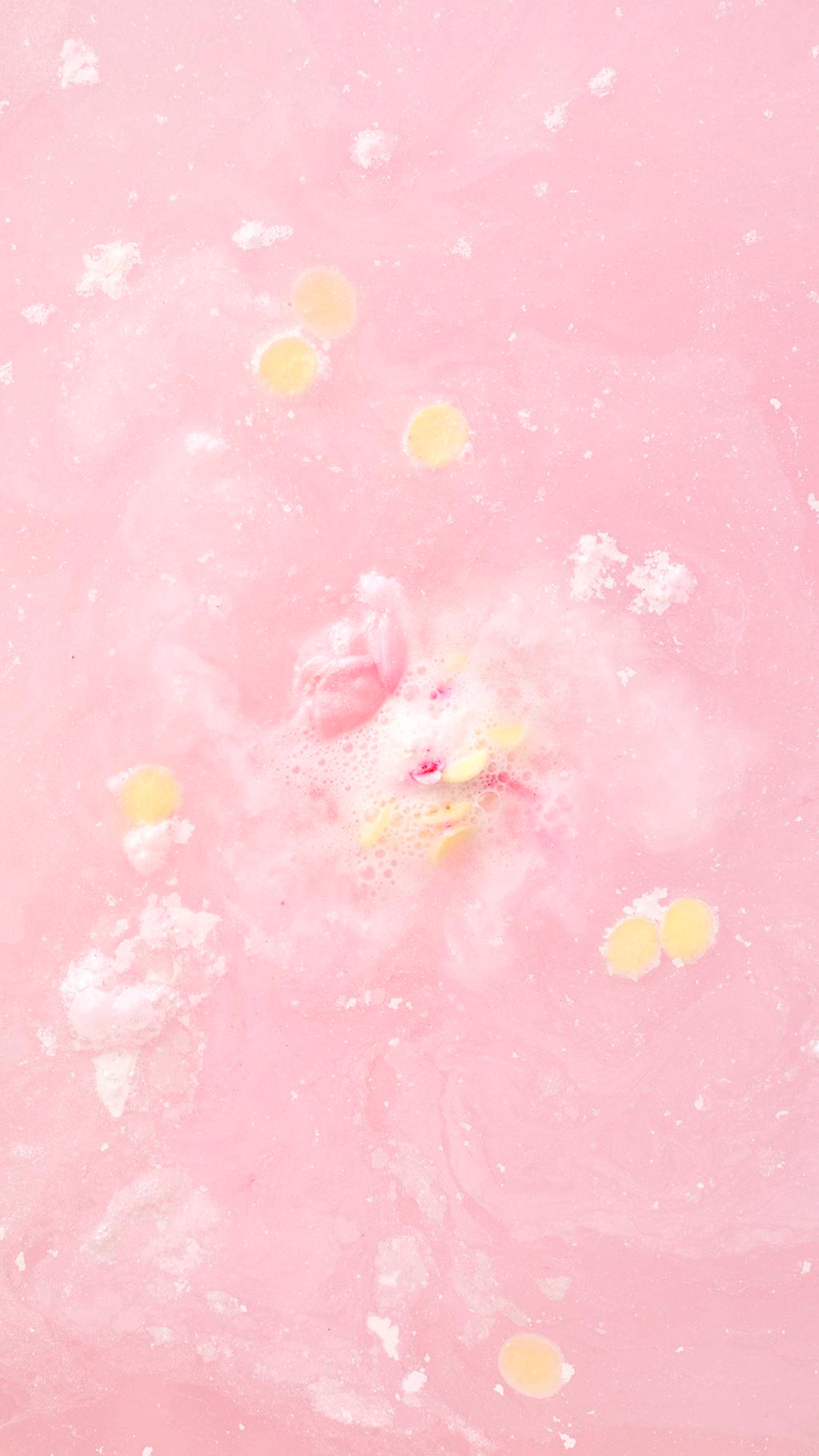 A top-down view of the Wedding bath bomb dissolved in pale pink water. A small pink rosebud and several tiny yellow circular petals float amidst white swirls and soft bubbles on the water's surface.