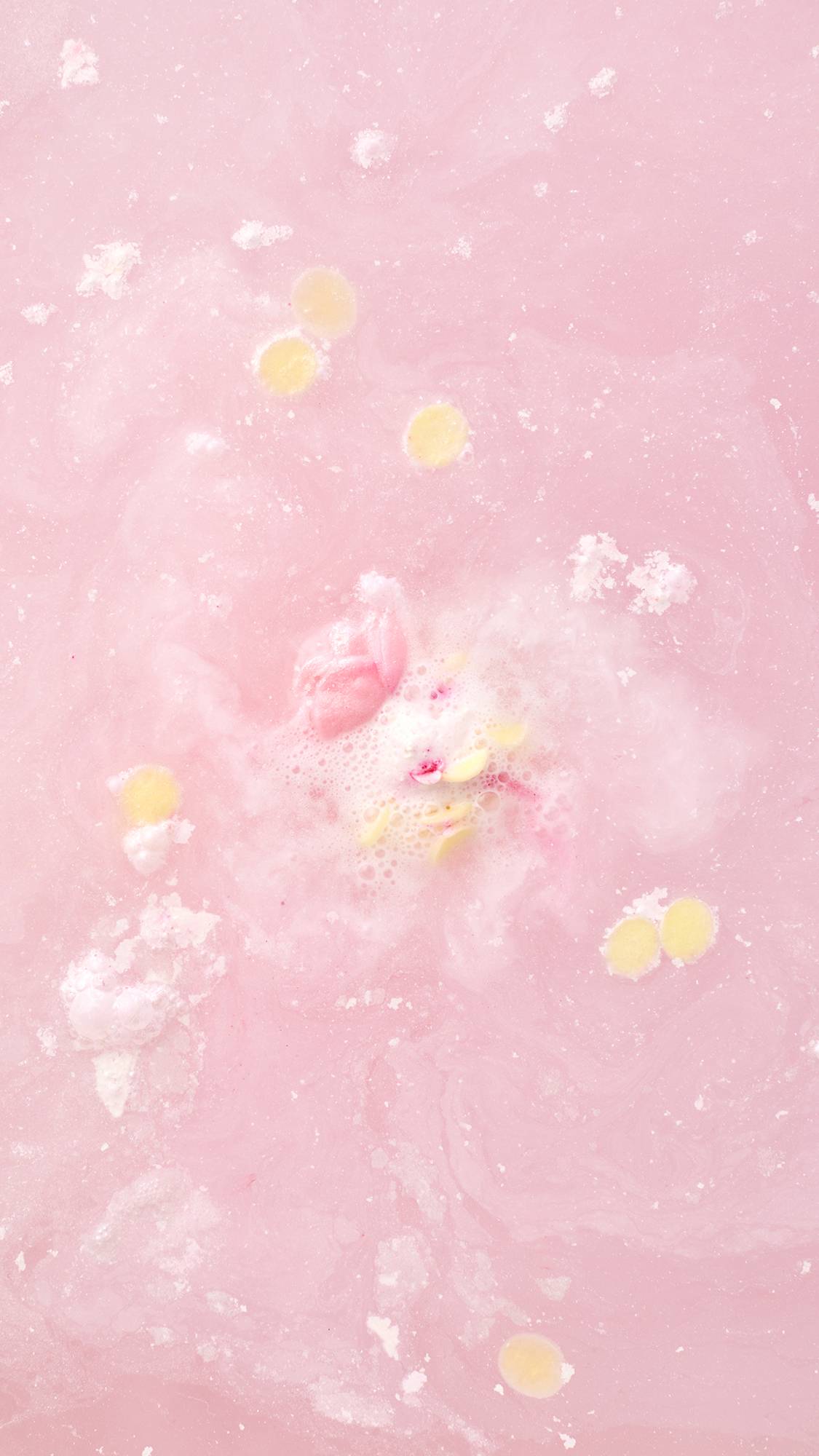 A top-down view of the Wedding bath bomb dissolved in pale pink water. A small pink rosebud and several tiny yellow circular petals float amidst white swirls and soft bubbles on the water's surface.