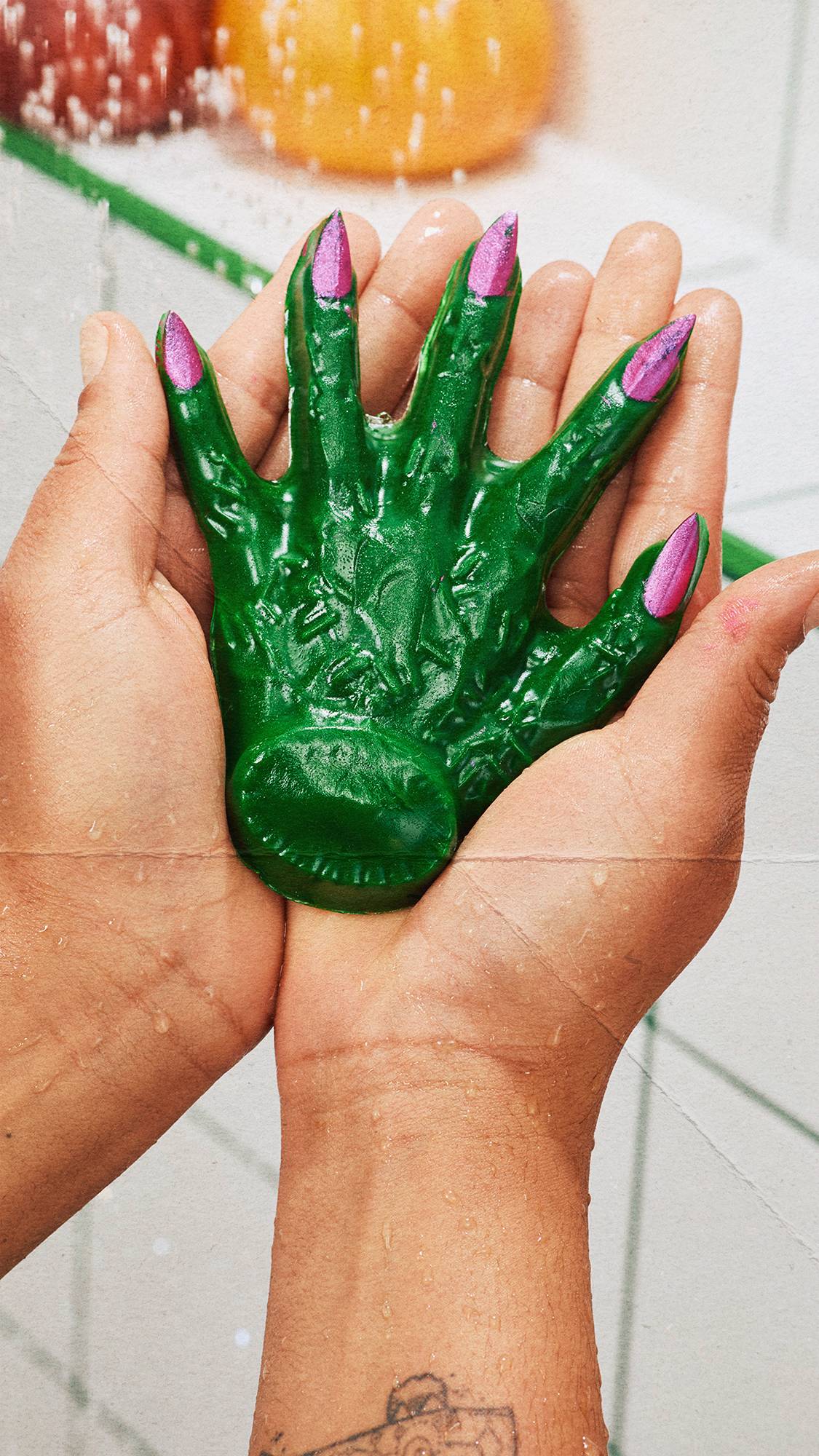 Two cupped hands hold a bright green, textured shower gummy shaped like a witch's hand with metallic pink fingernails. Water droplets dot the wet skin. White square tiles with green grout lines and faint pumpkins are in the background.