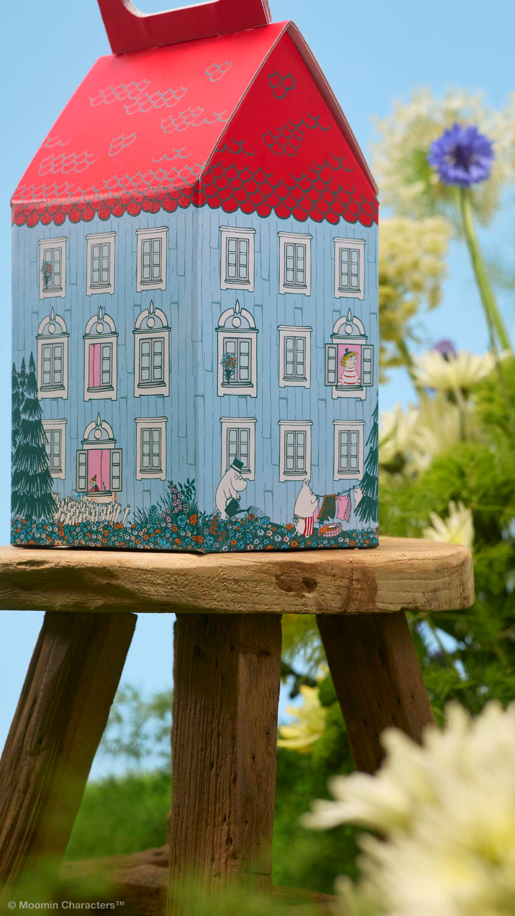 A close-up shot of a house-shaped gift box resting on a rustic wooden stool. The blue box features detailed illustrations of Moomin characters in the garden and windows. Out-of-focus green foliage and wildflowers surround it under a blue sky.