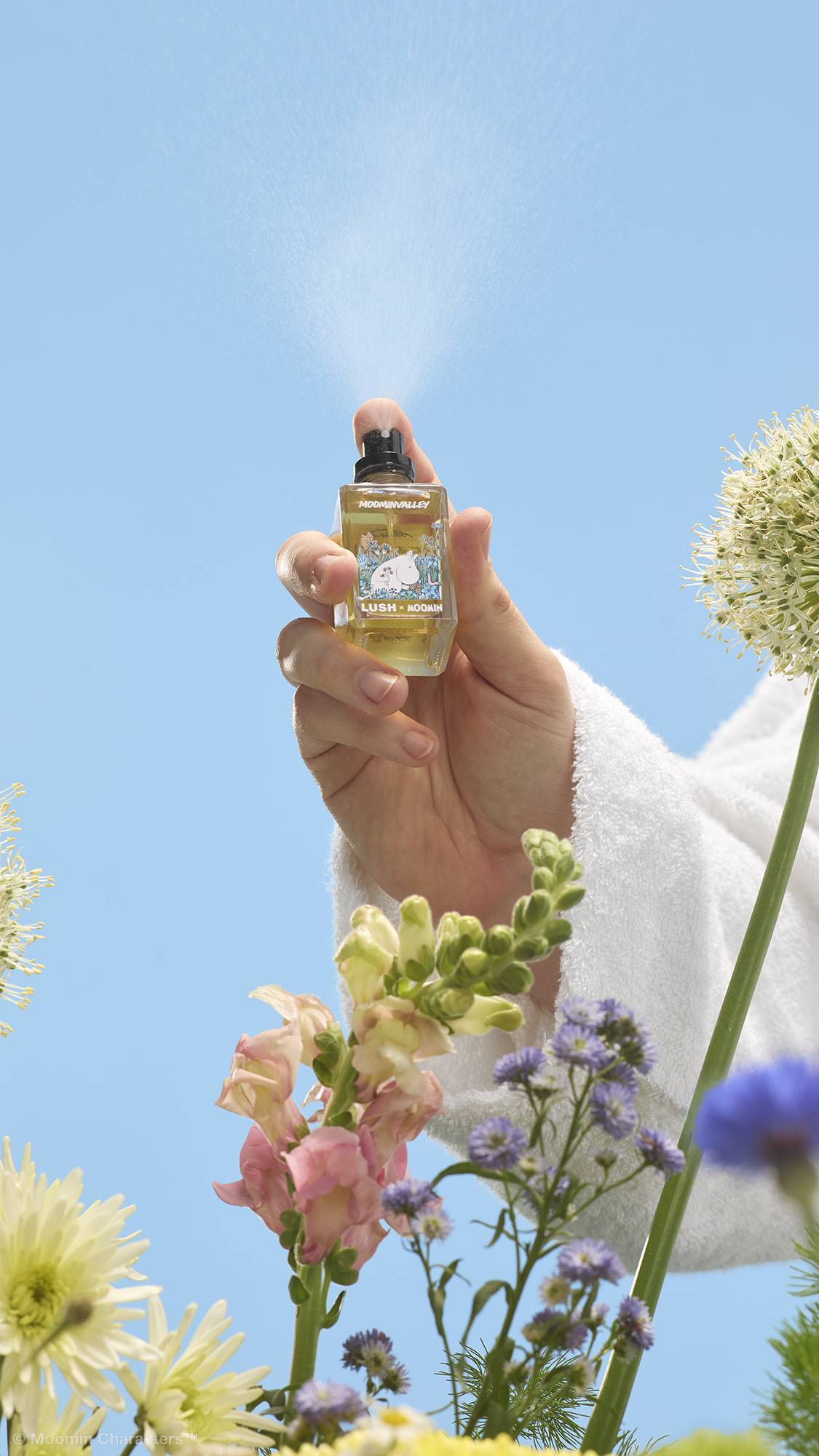 A close-up shot of a hand in a white dressing gown pressing the nozzle of a Lush x Moomin perfume bottle, releasing a fine mist into the air. Wildflowers are visible in the foreground against a clear blue sky.