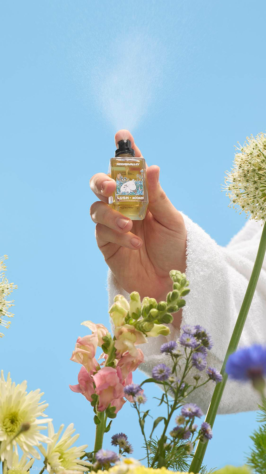 A close-up shot of a hand in a white dressing gown pressing the nozzle of a Lush x Moomin perfume bottle, releasing a fine mist into the air. Wildflowers are visible in the foreground against a clear blue sky.