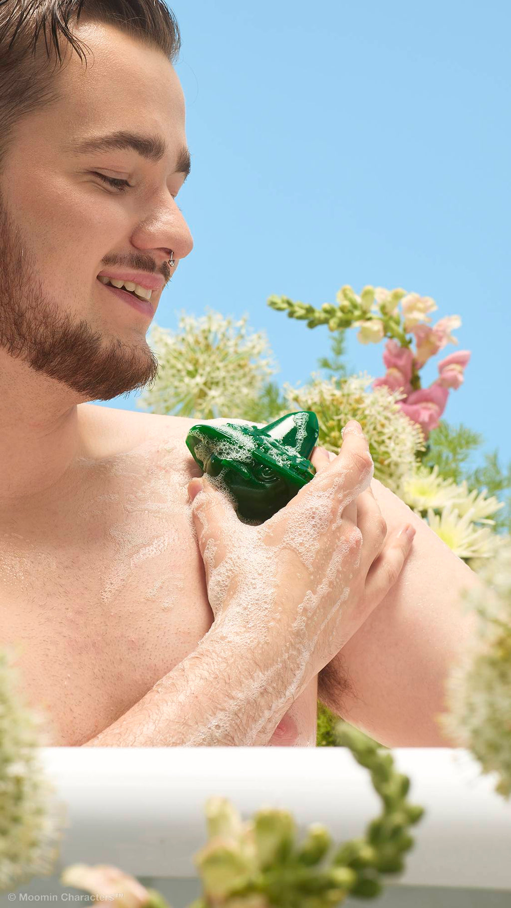 A close-up shot of a smiling person with a beard in a white bathtub, using a green Snufkin-shaped shower gummy to lather their shoulder. White and pink wildflowers are visible in the background against a clear blue sky.