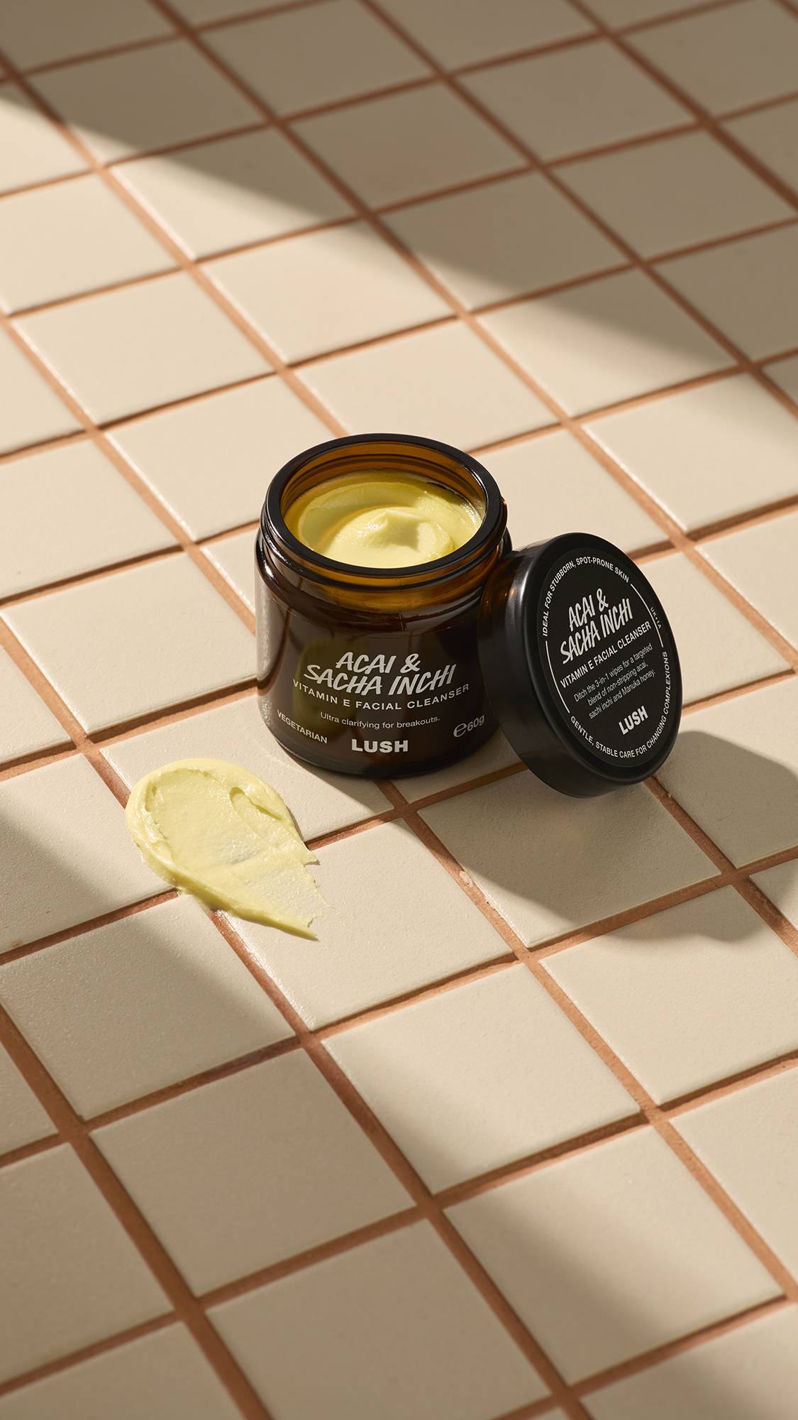 An open brown glass jar of Lush "Acai & Sacha Inchi" Vitamin E facial cleanser sits on a beige tiled surface. A thick dab of the pale yellow, creamy product is smeared on the tiles next to the jar and its black lid.