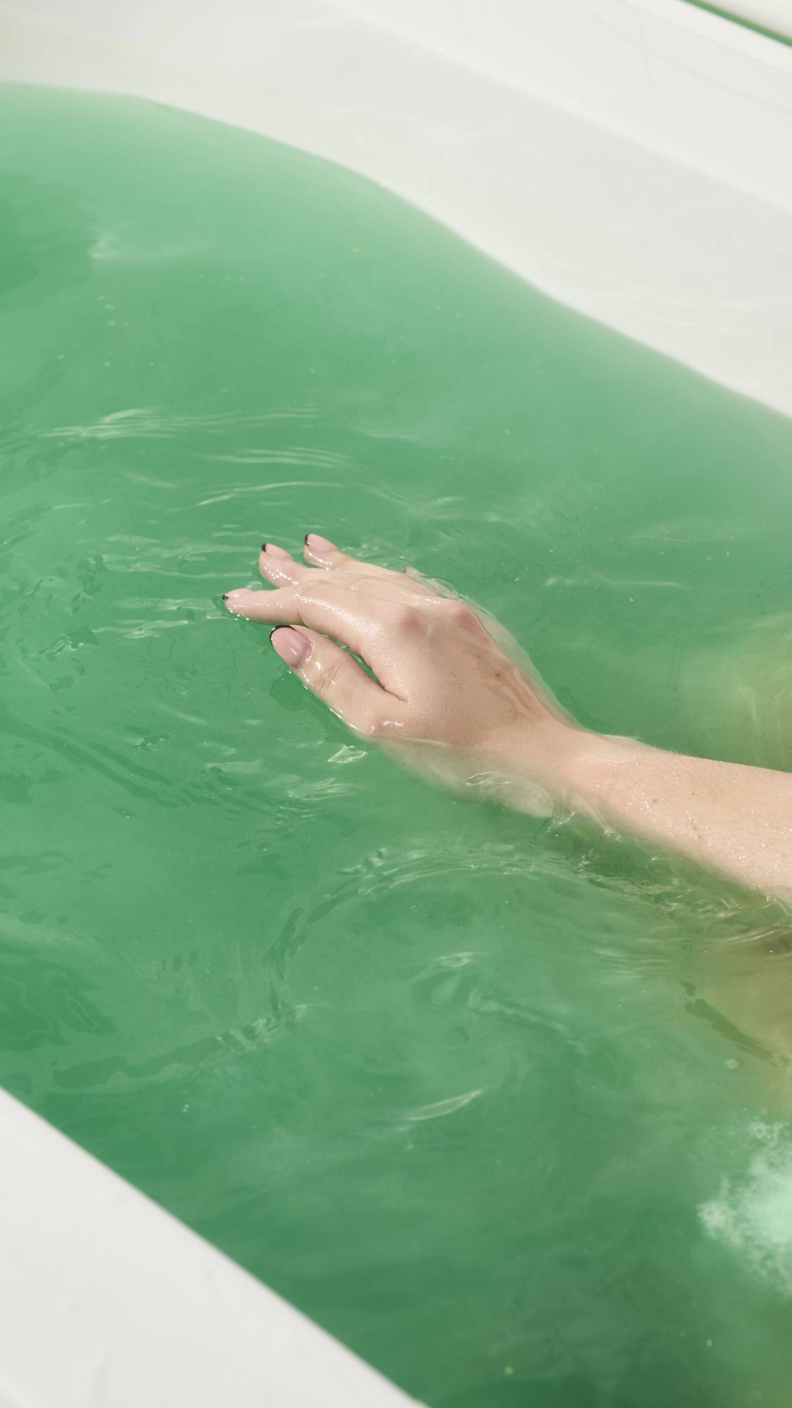 A close-up view of a hand with a pink and black tip manicure resting gently on the surface of vibrant, turquoise-green bathwater. The white tub's edge is visible.