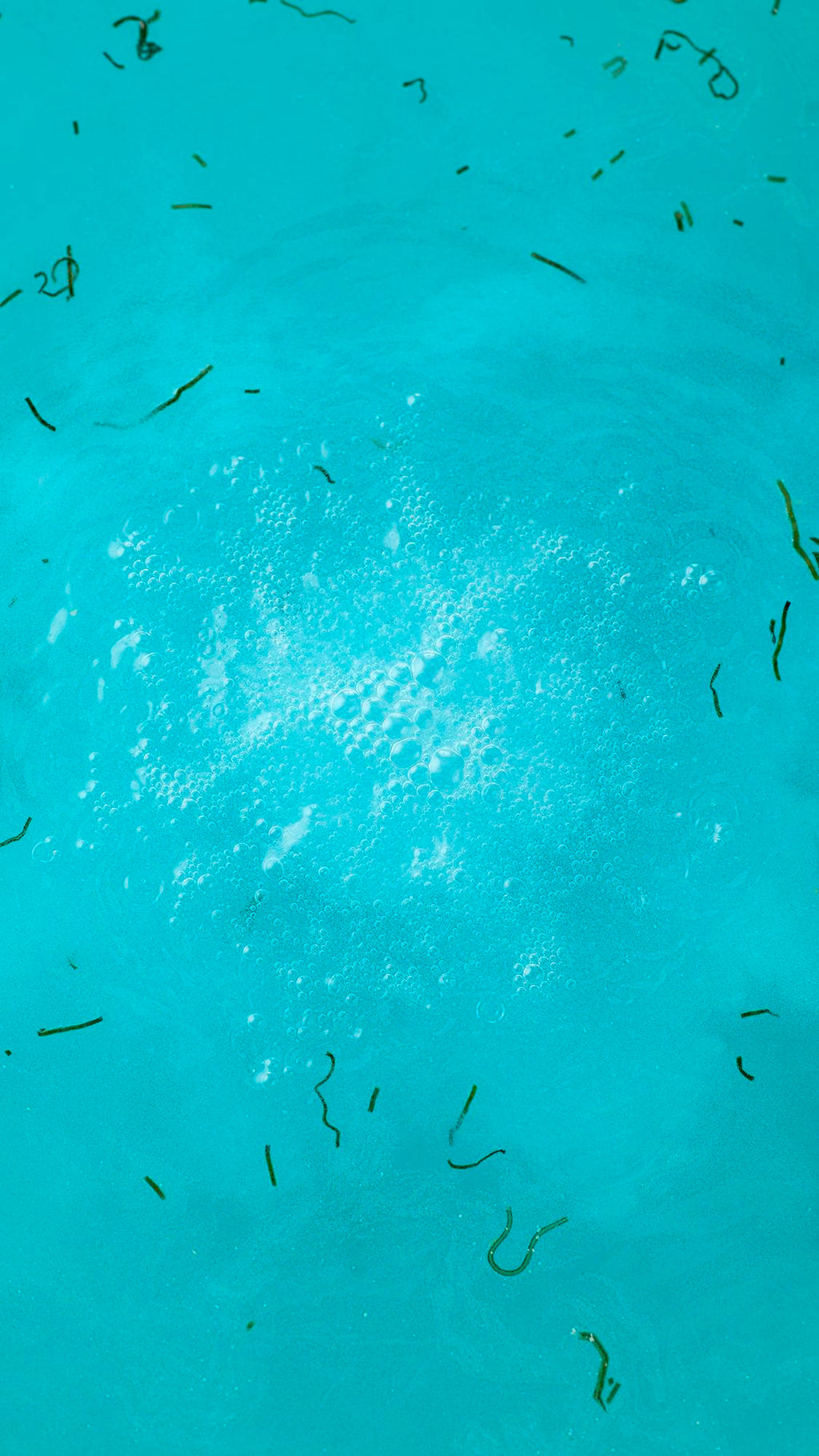 A top-down view of the dissolved Big Blue bath bomb in turquoise water. A frothy white cloud of bubbles sits in the center, surrounded by floating strands of dark green seaweed.