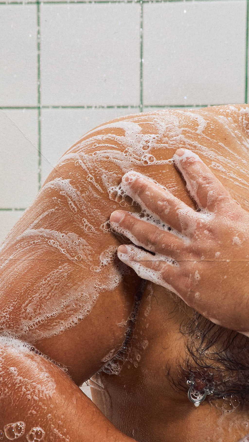 A close-up shot of a person lathering their soapy shoulder in the shower after using the Lush "BOO!" shower gel. A hand rubs thick, bubbly white foam onto wet skin, leaving frothy trails. White square wall tiles with green grout form the background.