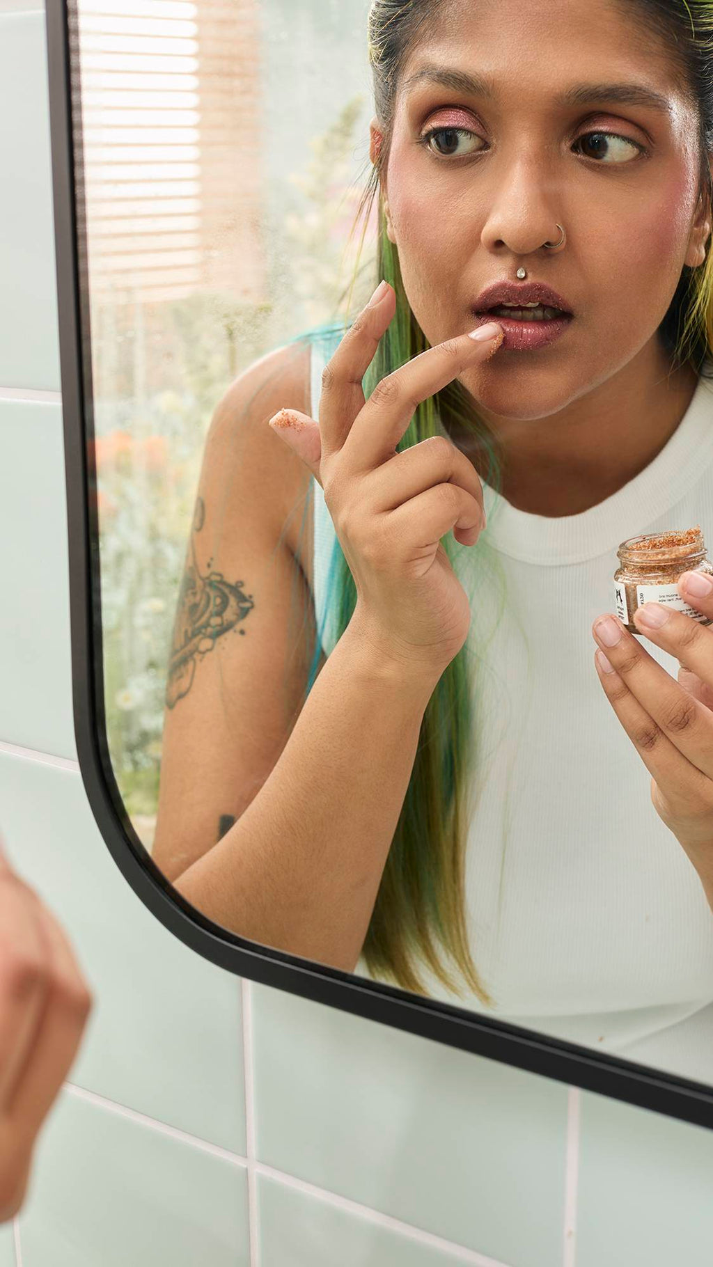A model with long green hair looks into a bathroom mirror while applying Lush "Buzz" Lip Scrub. They hold the small open jar in one hand and use their other index finger to gently rub the textured product onto their lower lip.