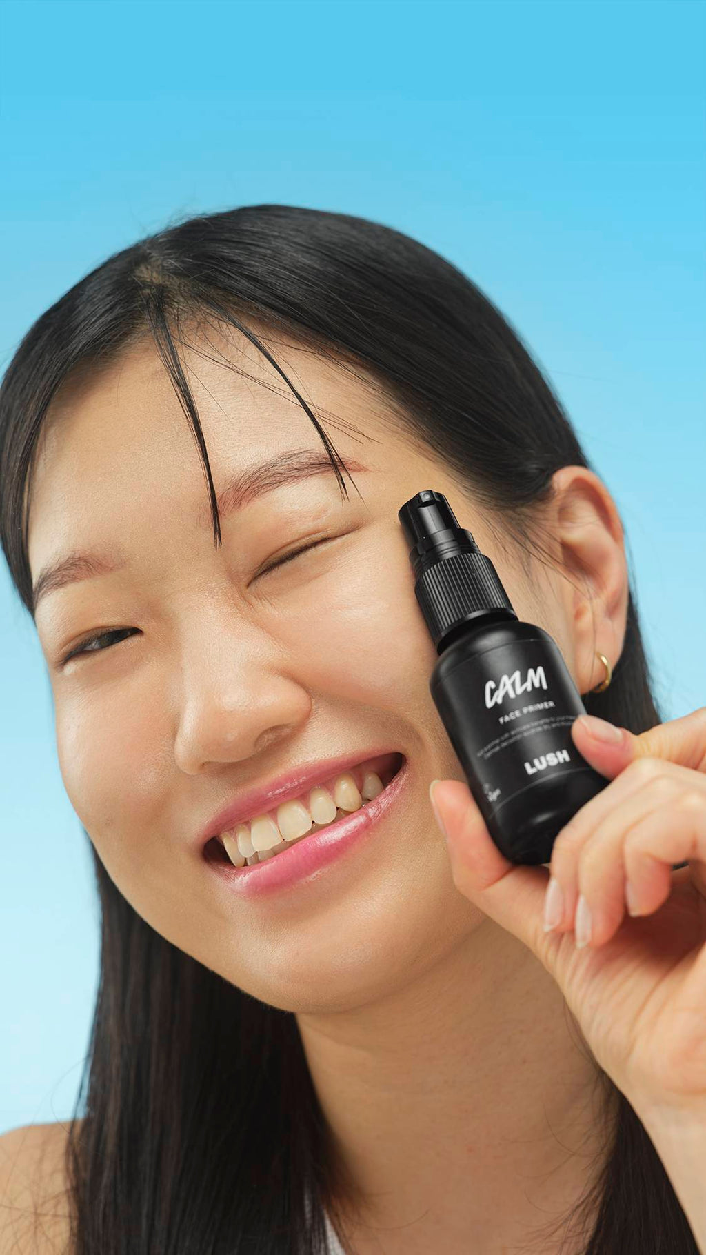 A close-up vertical portrait of a smiling person with dark hair, winking while holding the small black bottle of LUSH Calm Face Primer up to their cheek. The background is a bright blue.