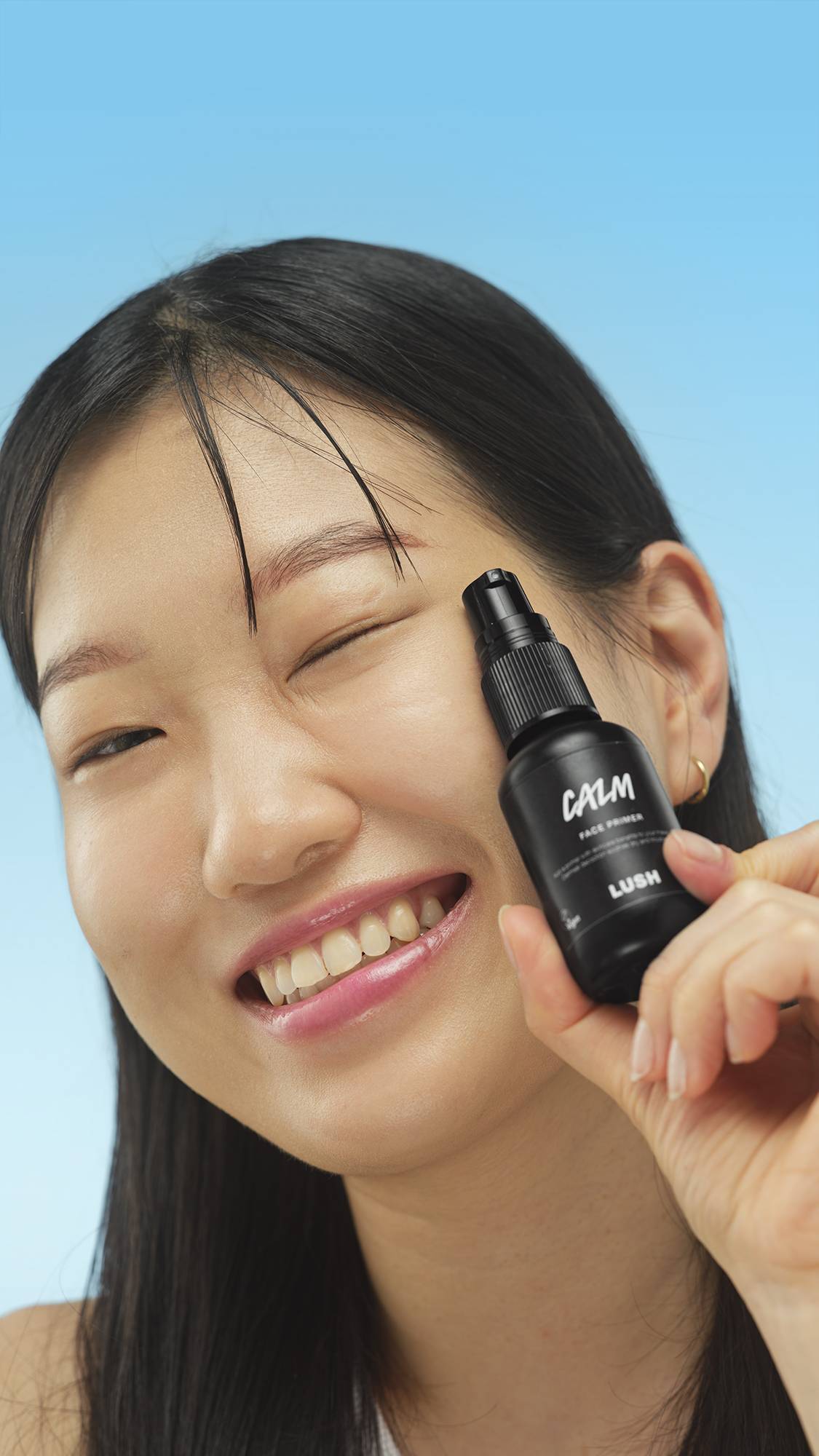 A close-up vertical portrait of a smiling person with dark hair, winking while holding the small black bottle of LUSH Calm Face Primer up to their cheek. The background is a bright blue.