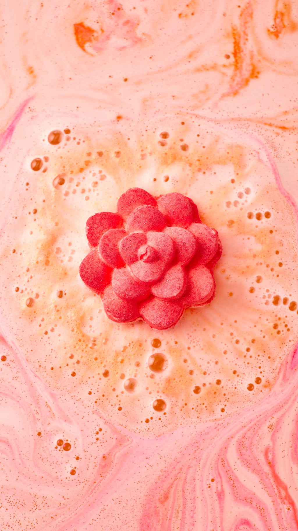 An aerial image of the "Comforter Rose' bath bomb as it begins to dissolve. It is surrounded by a ring of thick, cream-coloured bubbles, with swirling patterns of soft pink and peach radiating across the water's surface.