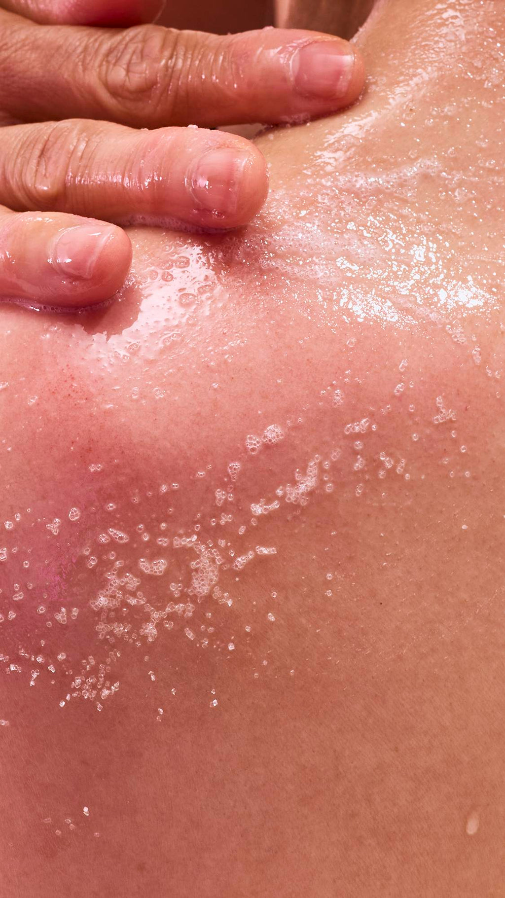 A close-up shot of wet skin with droplets of water and a trail of white, salt-like exfoliating granules. A hand with a pinkish tint is seen at the top, pressing down on the skin as if applying a scrub.