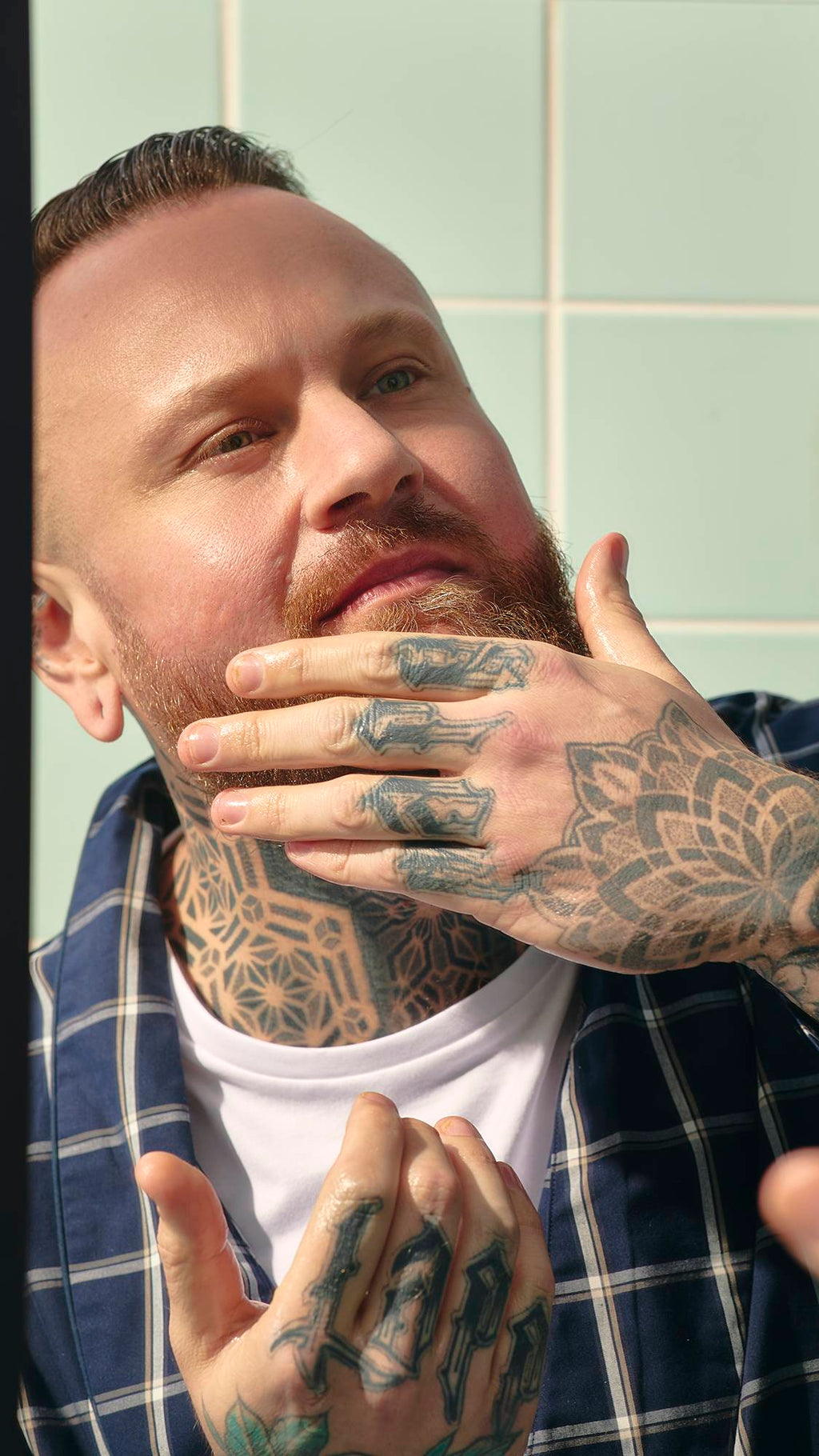 A model with a ginger beard and a blue-and-white checked dressing gown looks into a bathroom mirror. They have extensive tattoos on their neck and hands, and are using their fingers to massage the "Dirty" Hair and beard oil in.