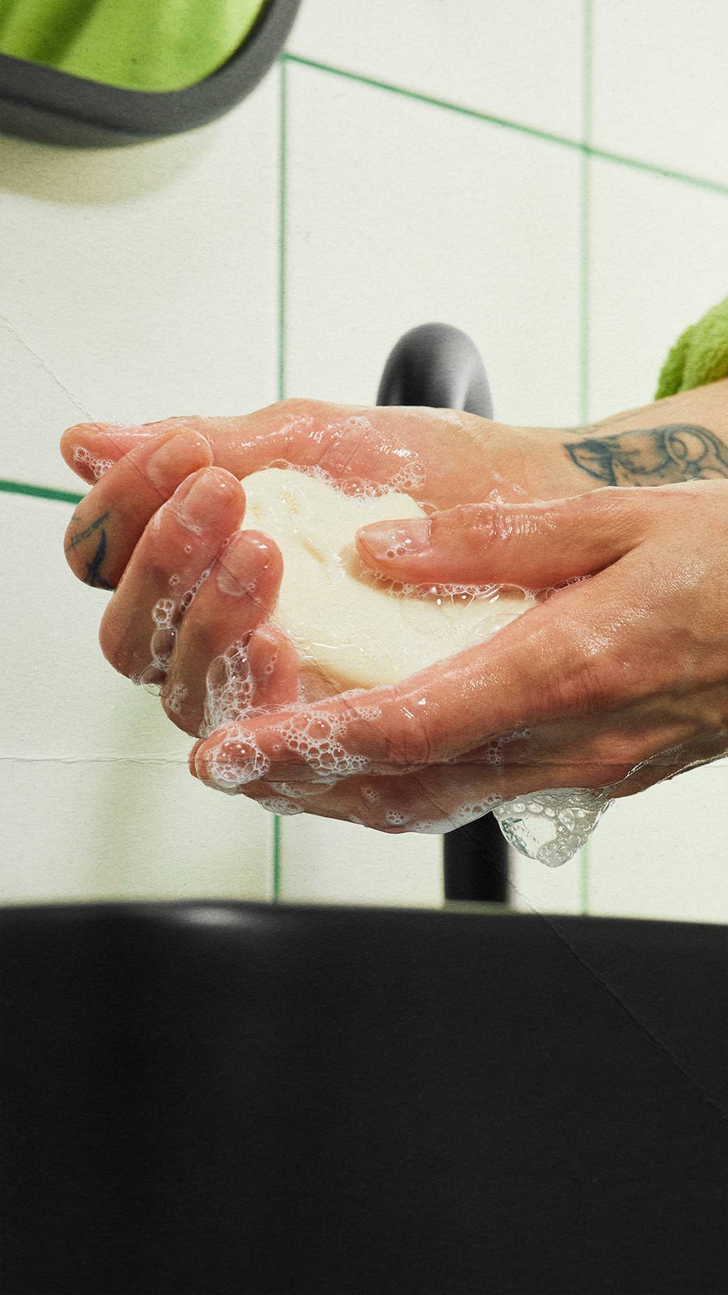 Tattooed hands lather an off-white, ghost-shaped bar of soap over a black sink, creating white soapy bubbles. A black tap and white wall tiles with green grout lines are visible in the background.