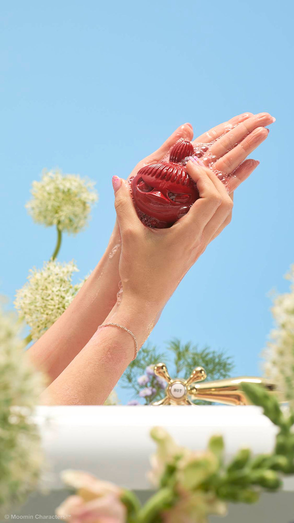 A close-up shot of two hands lathering a dark red Little My-shaped soap over the edge of a bathtub. Pink, frothy bubbles cover the hands, with a gold tap and wildflowers in the background against a clear blue sky.