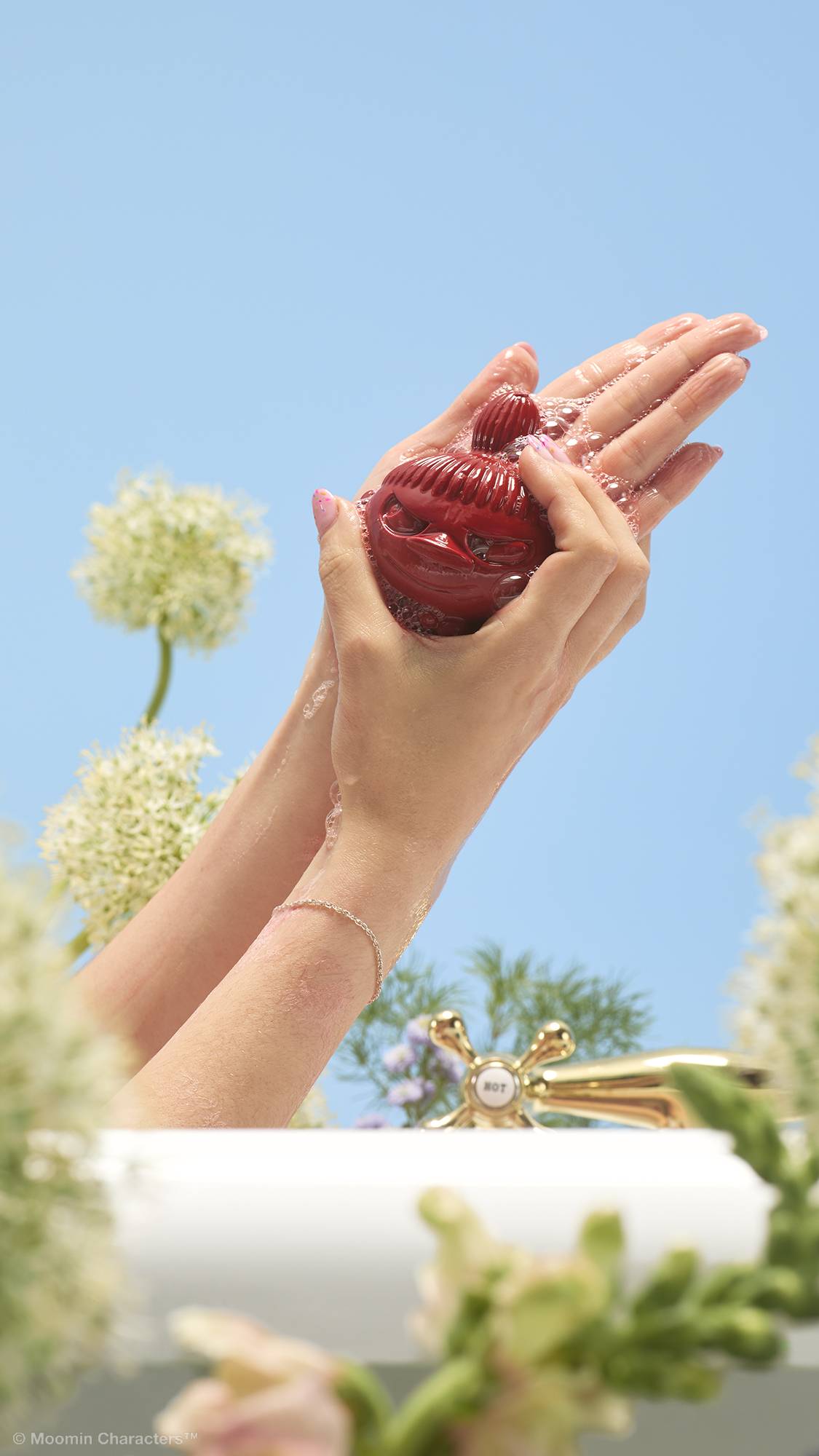 A close-up shot of two hands lathering a dark red Little My-shaped soap over the edge of a bathtub. Pink, frothy bubbles cover the hands, with a gold tap and wildflowers in the background against a clear blue sky.