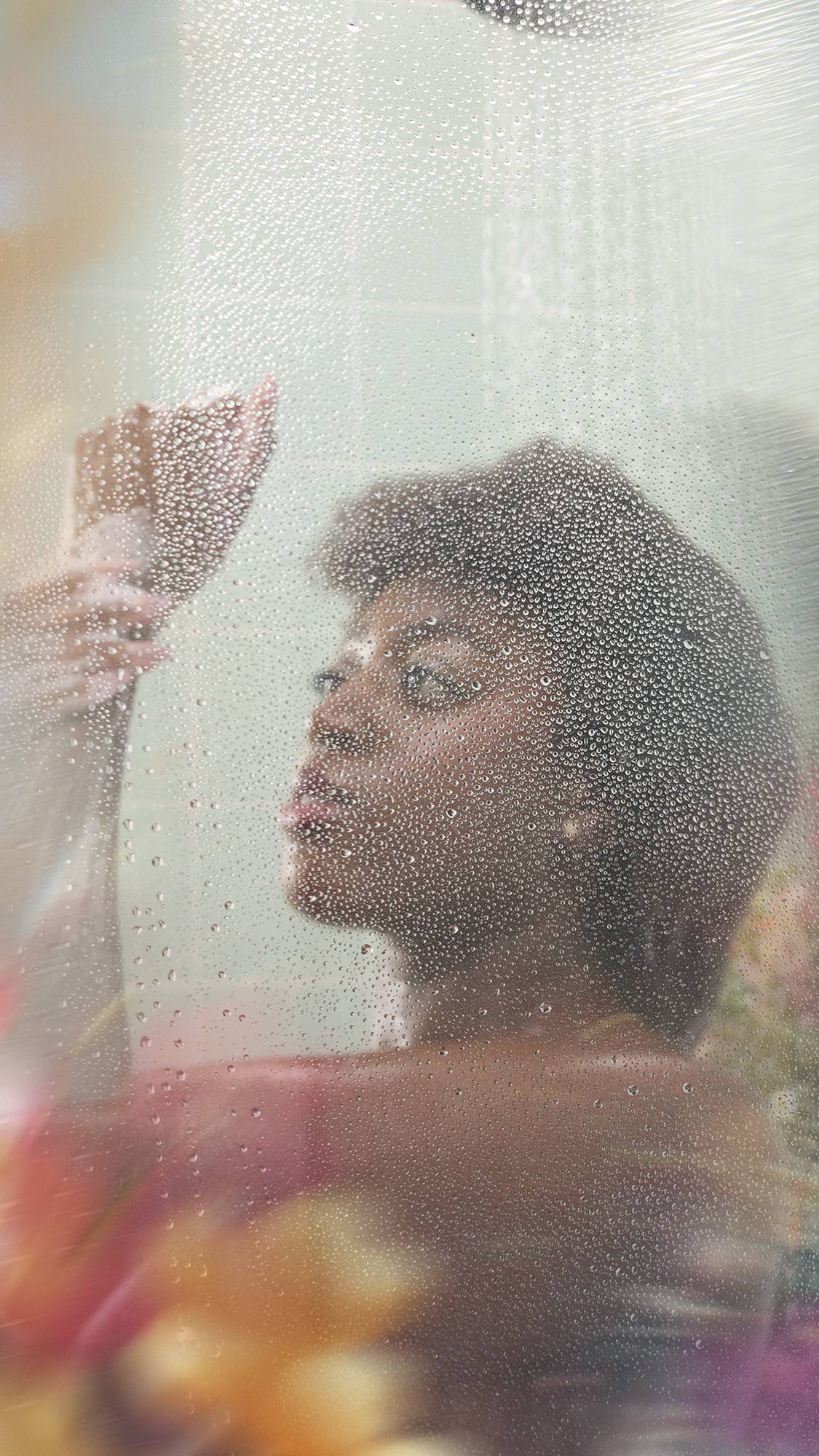 A person with short, dark curly hair stands behind a wet shower screen covered in water droplets. They are looking upwards with one hand raised near their face, set against a backdrop of mint green tiles and blurred colorful flowers.