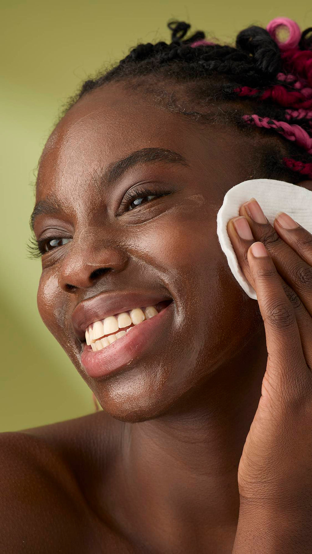 A woman with pink and black braids smiles as she removes the Oat facial cleanser from her face. The thick, creamy product stands out against her skin and the solid olive-green background, emphasising its rich texture.