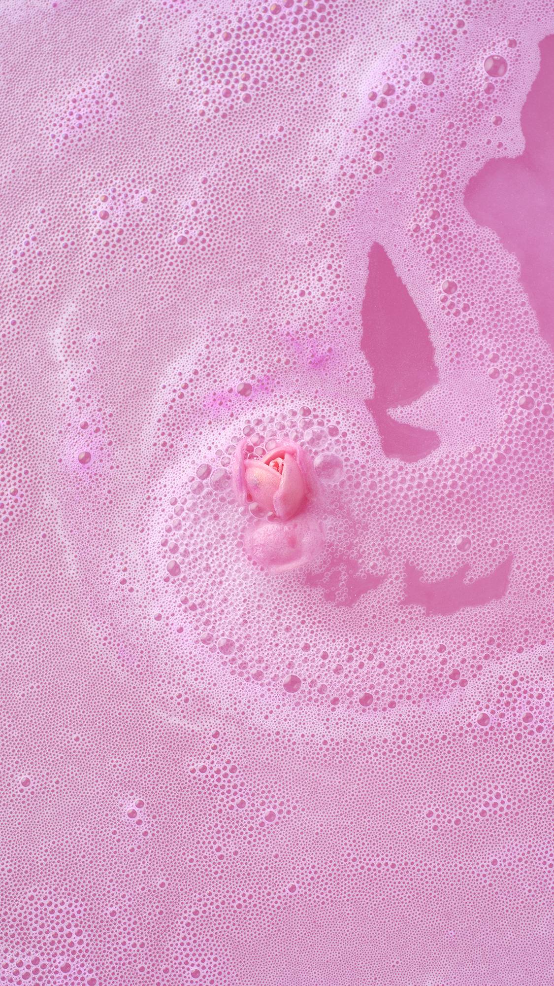 Top-down view of the Sex Bomb Bath Bomb fully dissolved, with its signature pink rosebud floating in the center of thick, frothy pink bathwater.