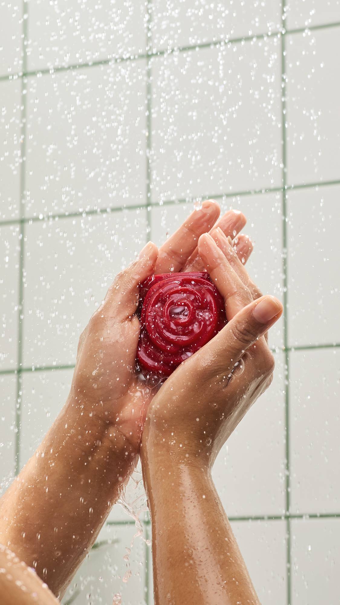 Two hands holding a red, rose-shaped "Sexy" shower gummy soap under spraying water, with water droplets splashing against a background of white tiles with green grout.