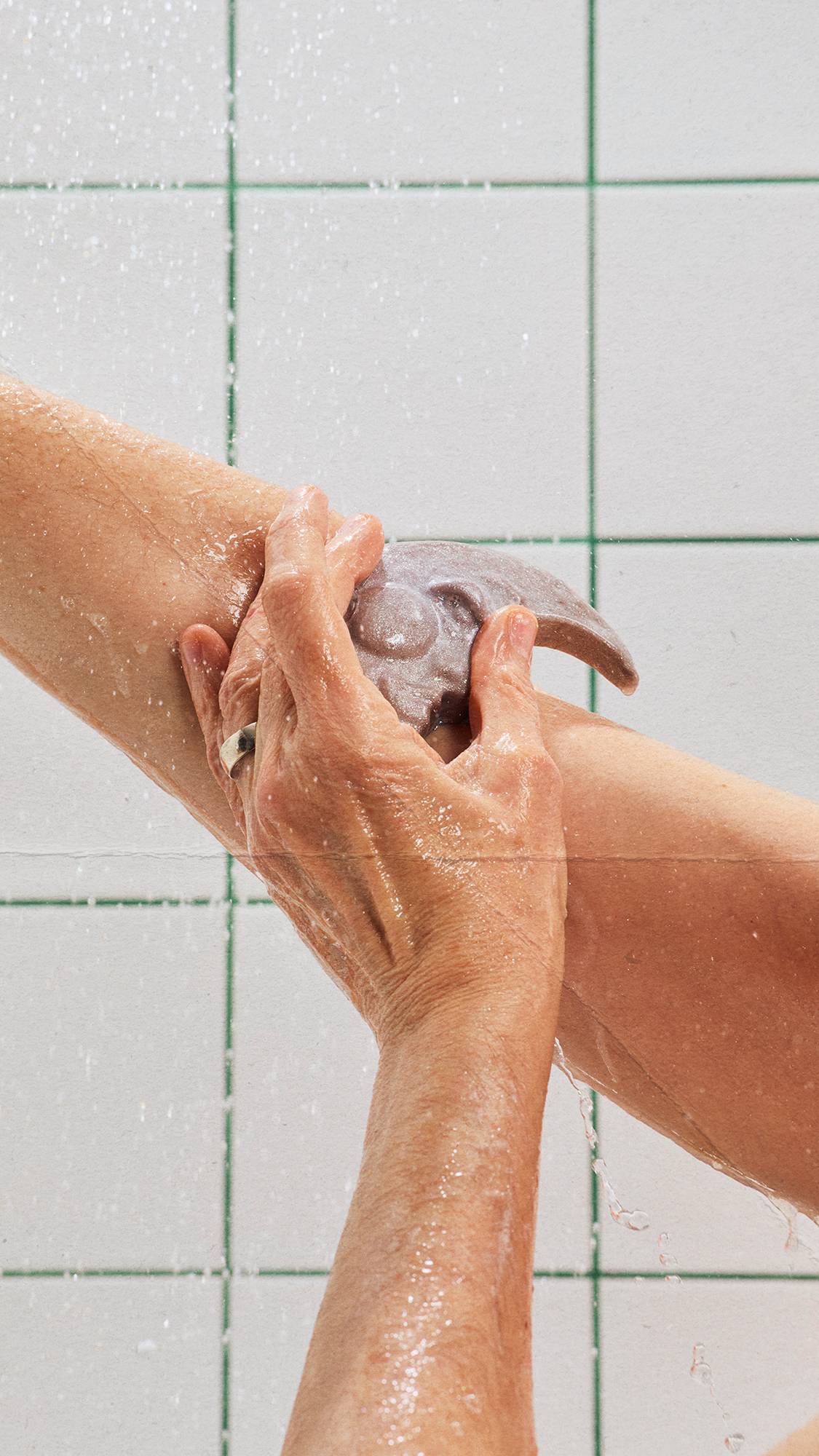The image shows wet hands lathering an arm with a silver, crescent moon-shaped soap bar in the shower. Water droplets spray across the scene against a background of white tiles with green grout lines.
