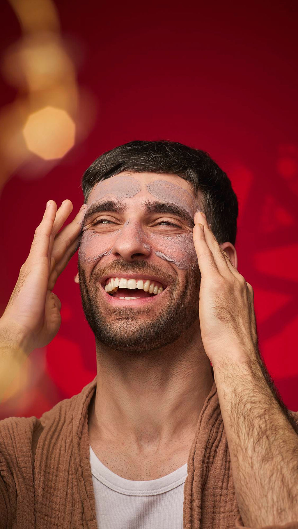 A person laughs while wearing the mauve-brown Sipak face mask across their forehead and cheeks. They wear a brown textured robe against a deep red background with soft golden bokeh lights.