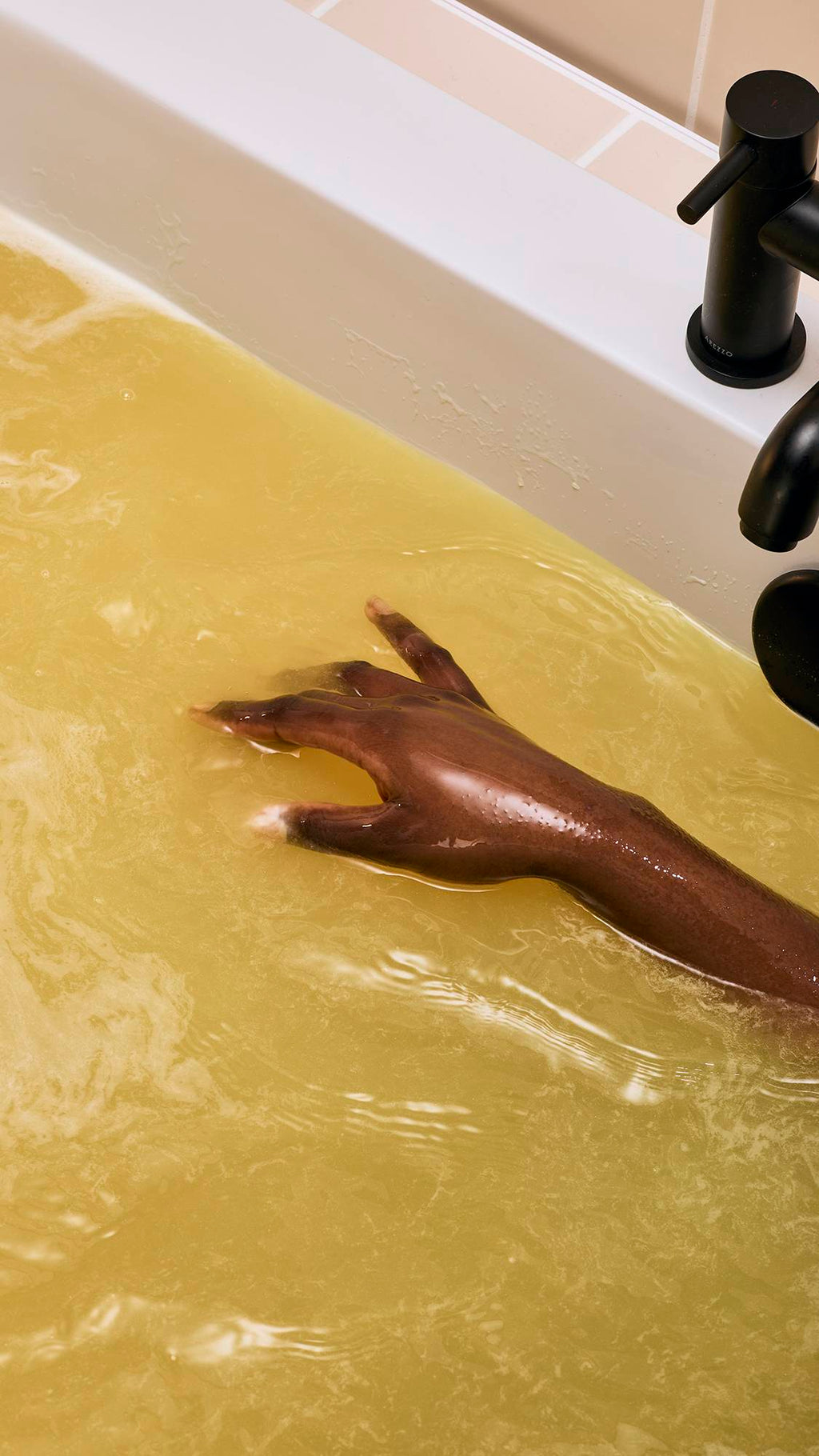A close-up of a model's hand gently swirling around a golden bath. Thick, creamy yellow foam swirls across the water's surface as a bath bomb dissolves, creating a marbled pattern of light and dark gold.