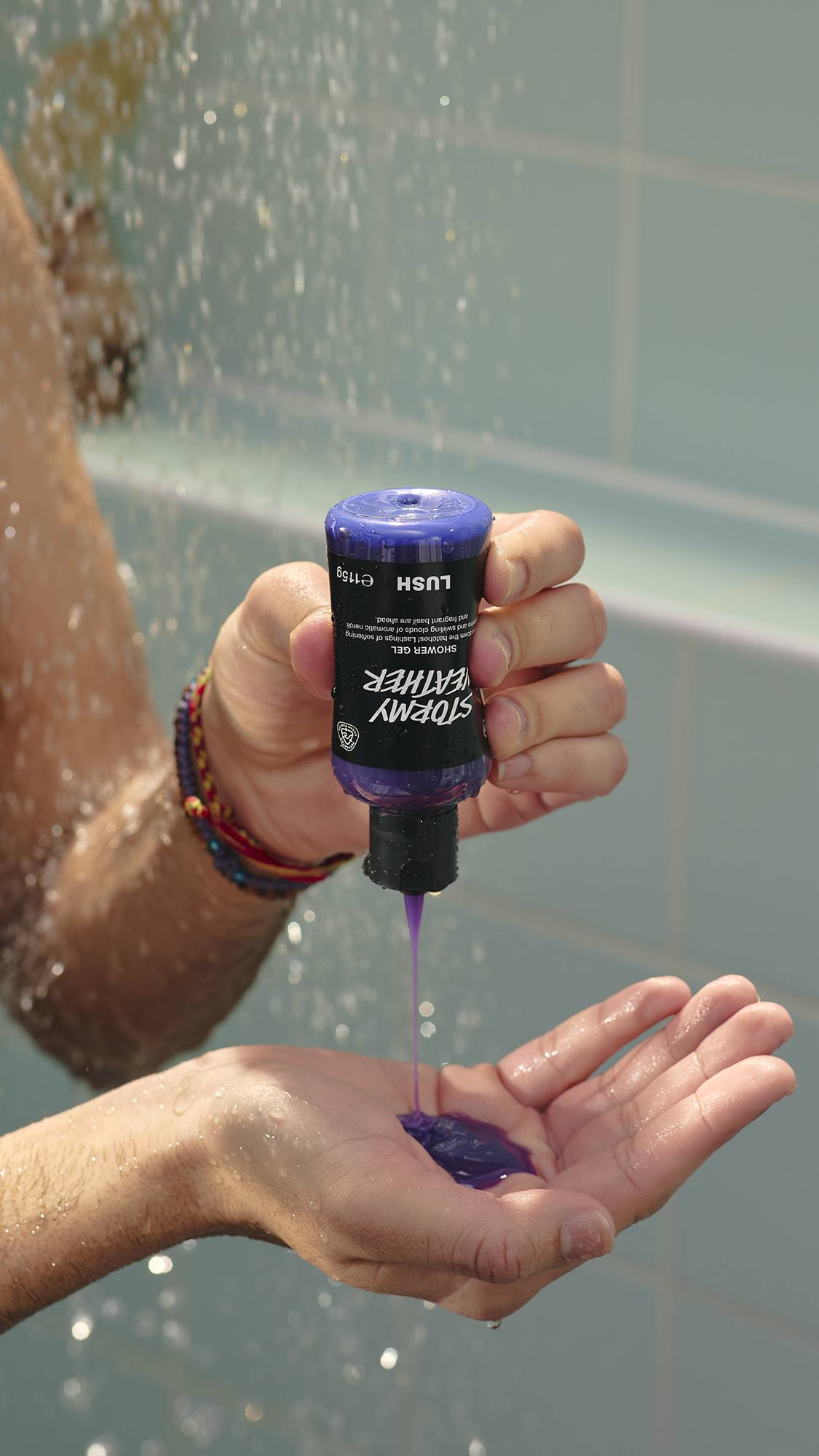 The image shows a model's hands holding a Lush bottle upside down, pouring thick, purple "Stormy Weather" shower gel into their open palm. The background shows a soft shower spray against light green bathroom tiles.