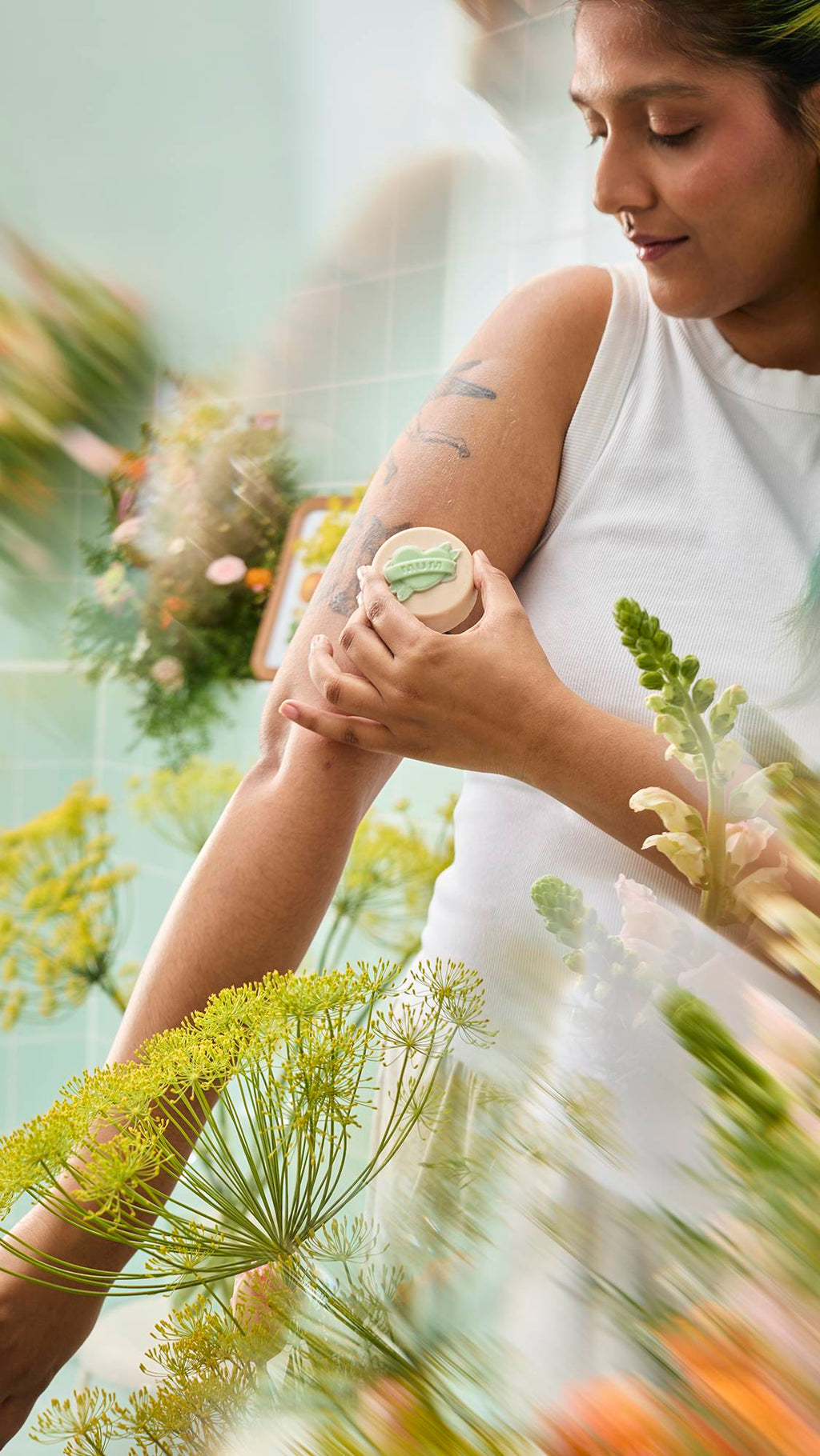A person wearing a white tank top applies a round, cream-coloured "Symbol of Love" body oil bar to their tattooed upper arm. The scene is framed by blurred yellow flowers against a background of mint green tiles.