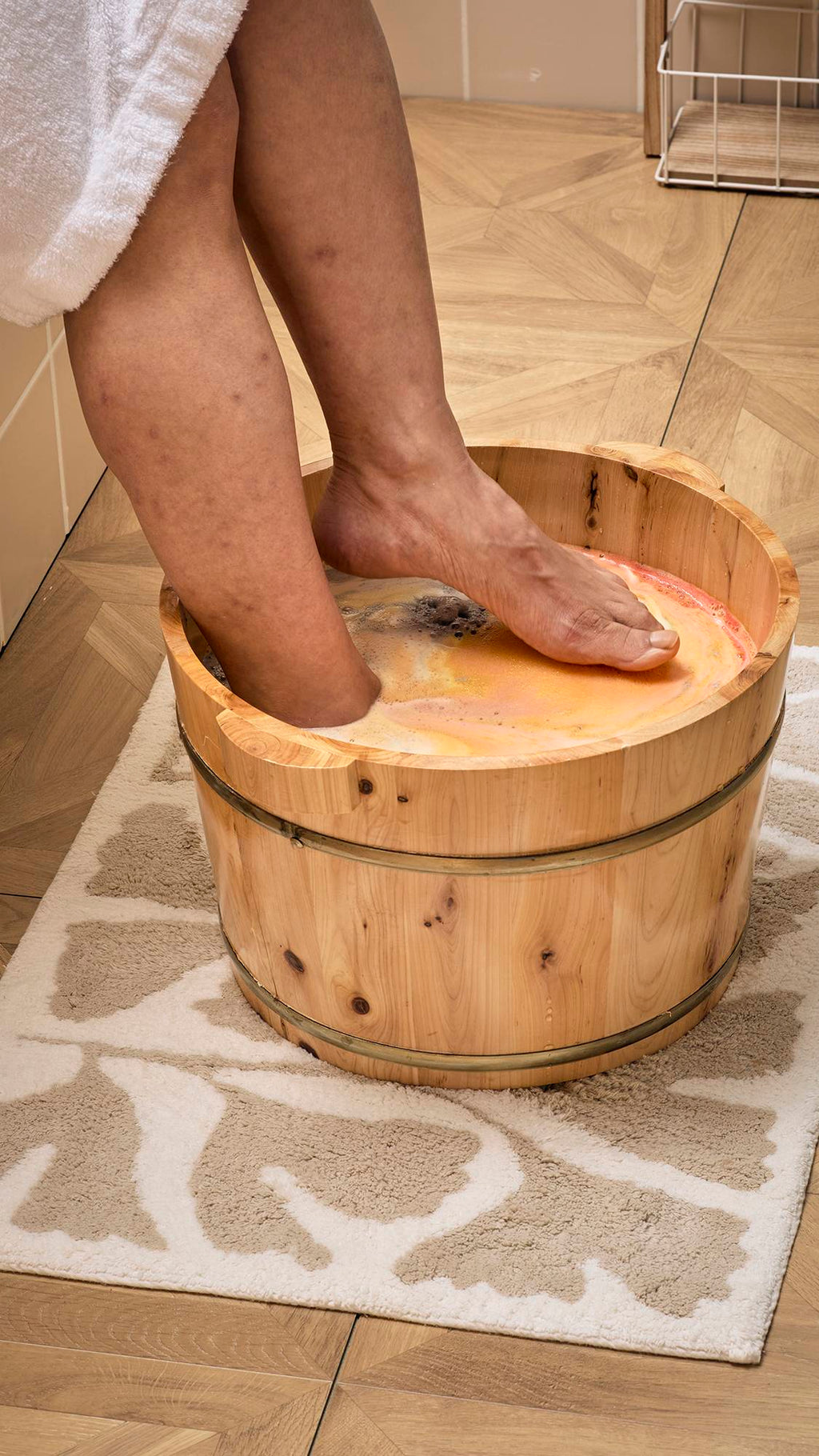 A model wearing a white dressing gown places their feet into a wooden half-barrel tub filled with orange-tinted water. Swirls of yellow, pink, and grey foam from the dissolving "Volcano" foot soak cover the surface of the foot spa.