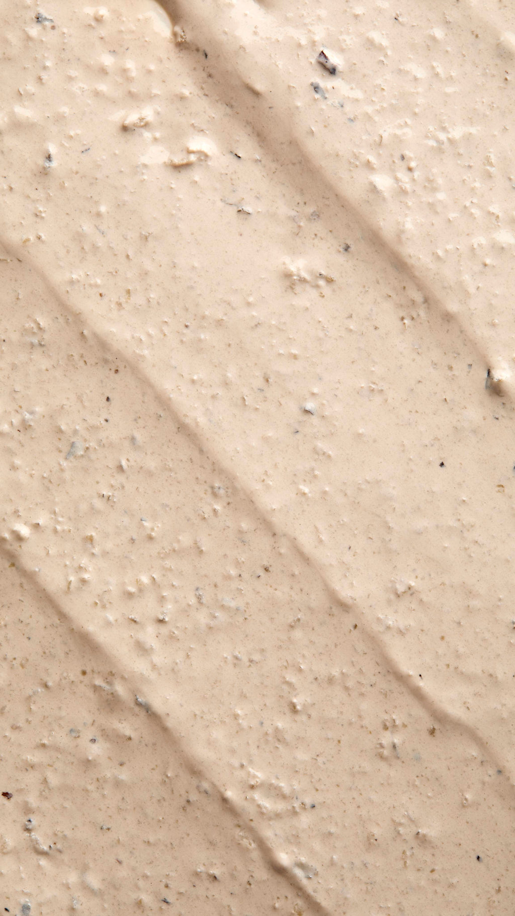 A close-up, textured shot of "Tiny Musicians Of The Forest, a creamy, beige-coloured face mask spread in thick, diagonal ridges. The surface is grainy and lumpy, containing small dark exfoliating particles and flecks.