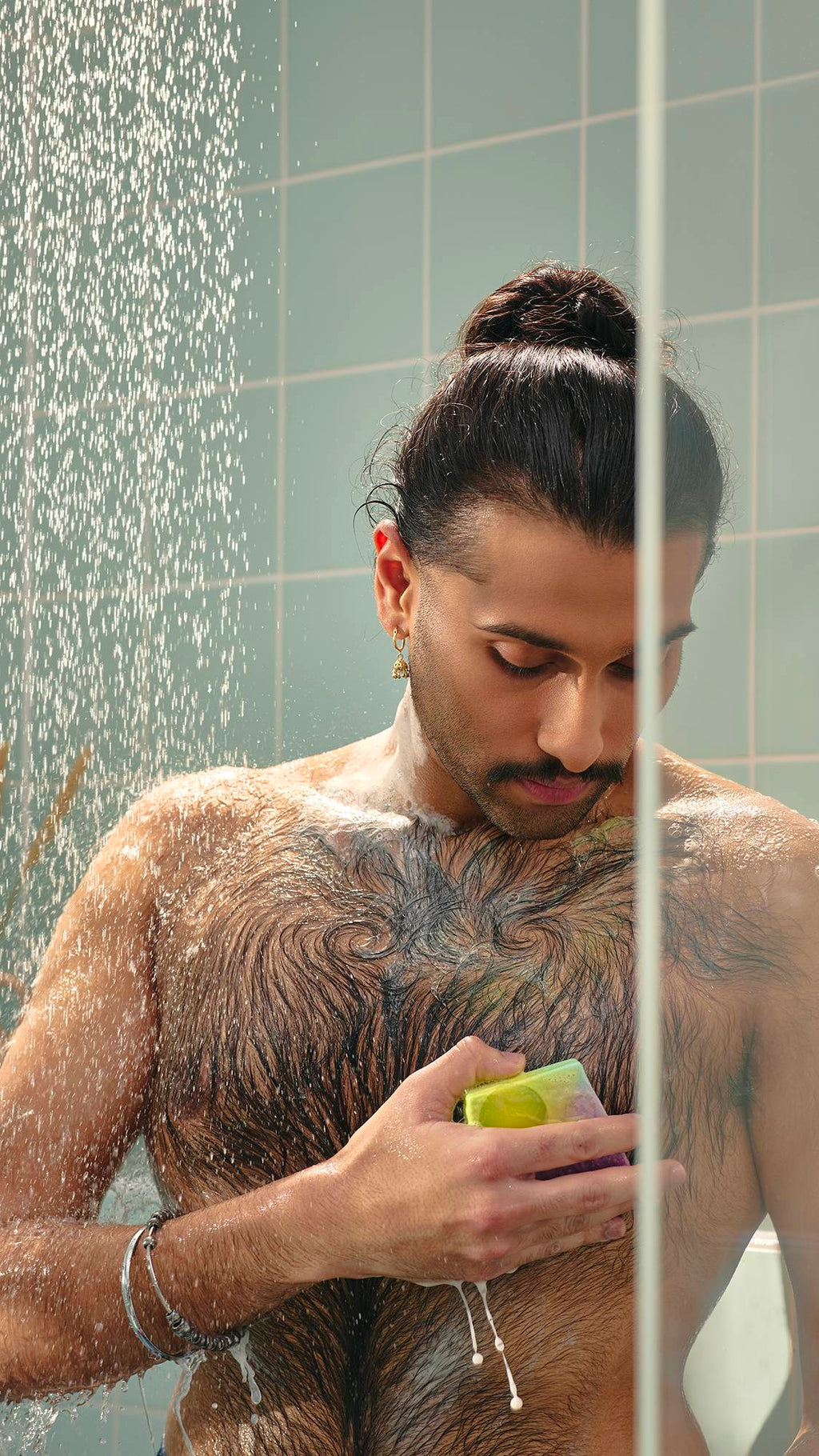 The image shows a model with dark hair in a bun looking down as they lather their chest with a slice of marbled purple and green "To The Moon and Back" soap in the shower. The water creates a fine mist around