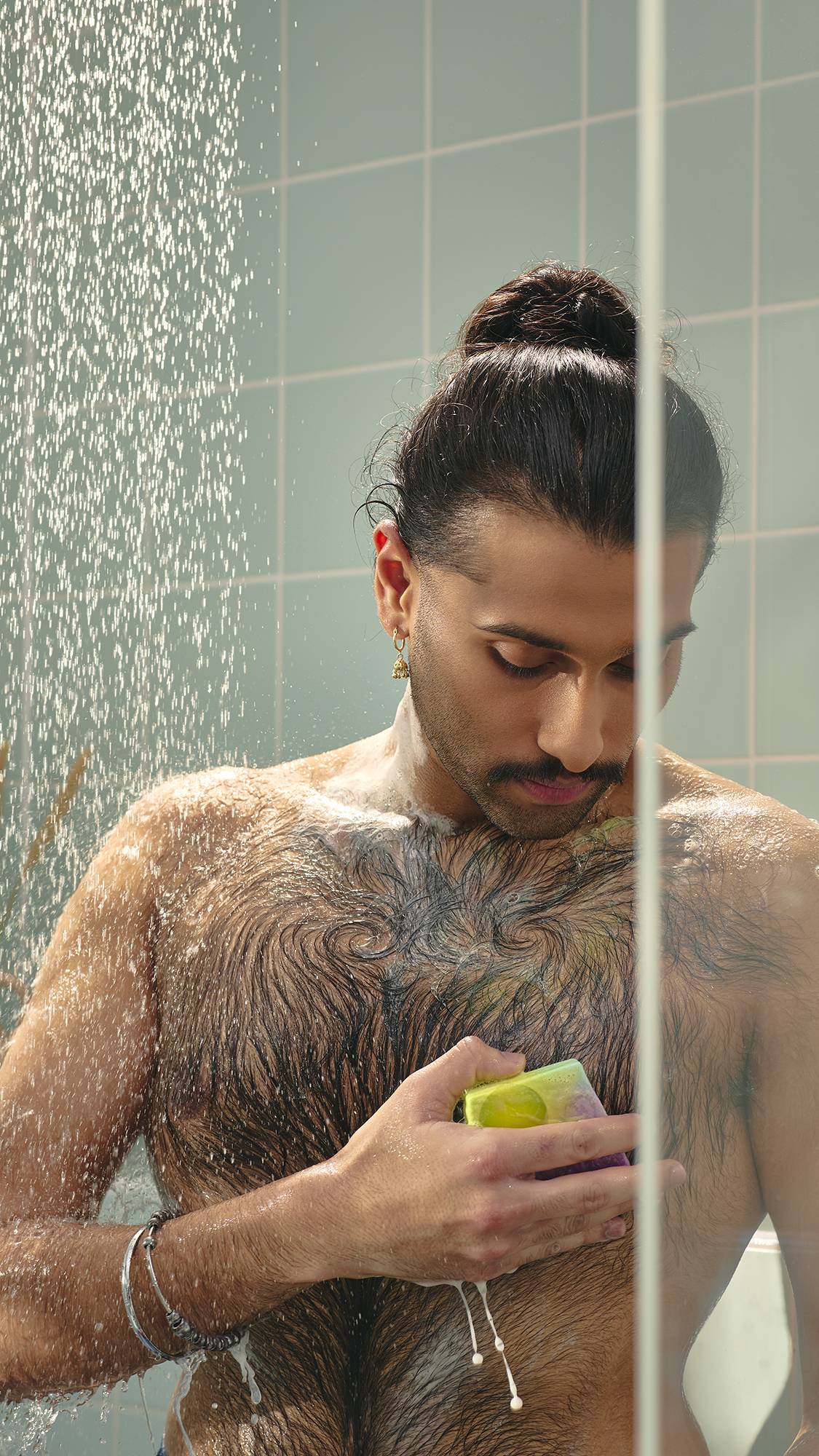 The image shows a model with dark hair in a bun looking down as they lather their chest with a slice of marbled purple and green "To The Moon and Back" soap in the shower. The water creates a fine mist around