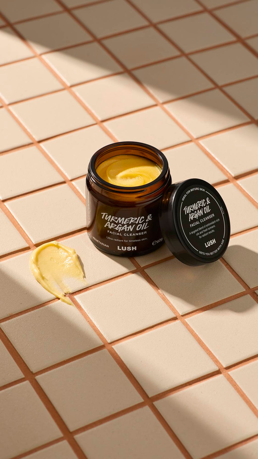 An open brown glass jar of Lush "Turmeric And Argan" facial cleanser sits on a beige tiled surface. A thick dab of the pale yellow, creamy product is smeared on the tiles next to the jar and its black lid.