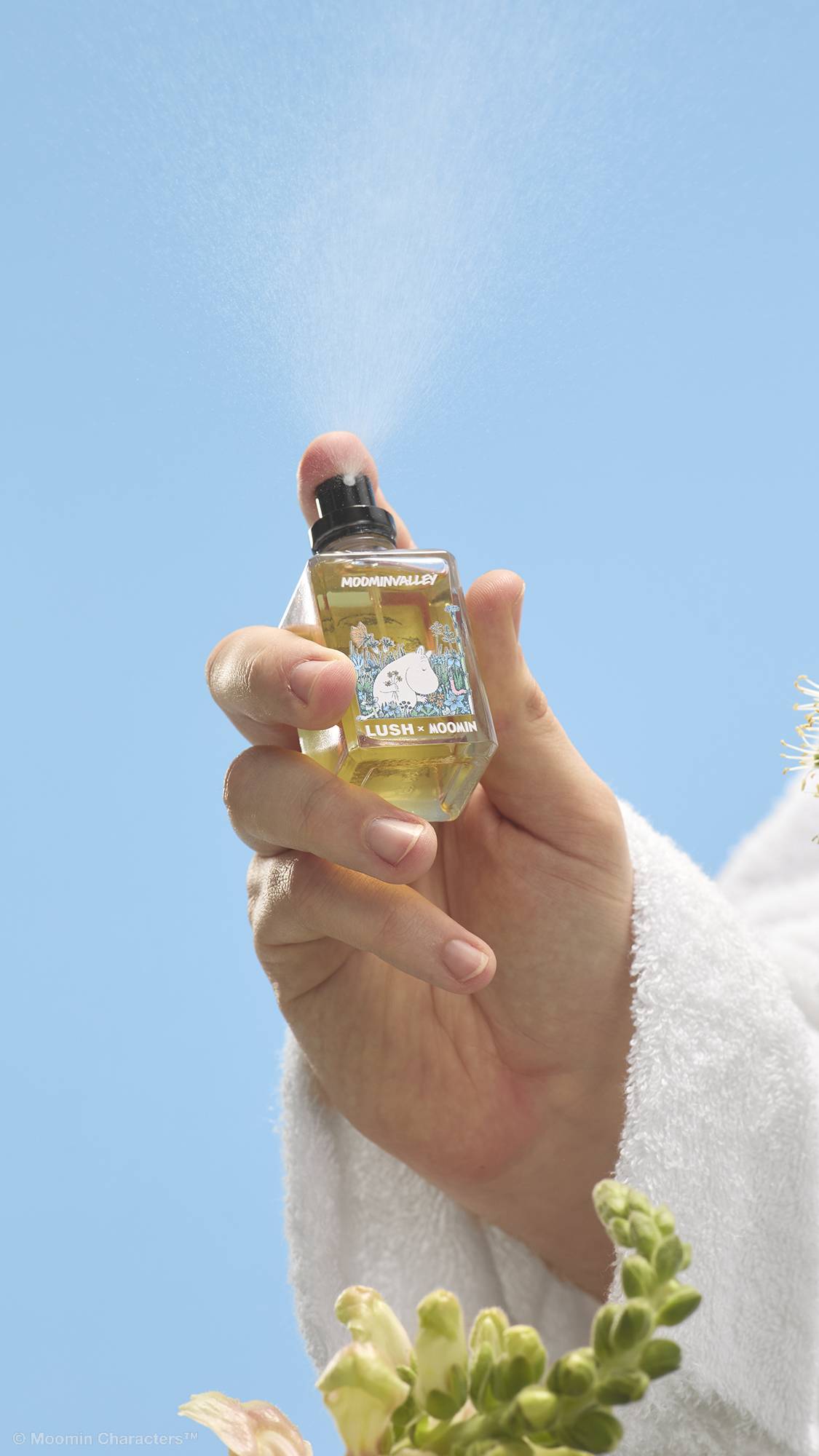 A close-up shot of a hand in a white dressing gown pressing the nozzle of a Lush x Moomin perfume bottle, releasing a fine mist into the air. Wildflowers are visible in the foreground against a clear blue sky.