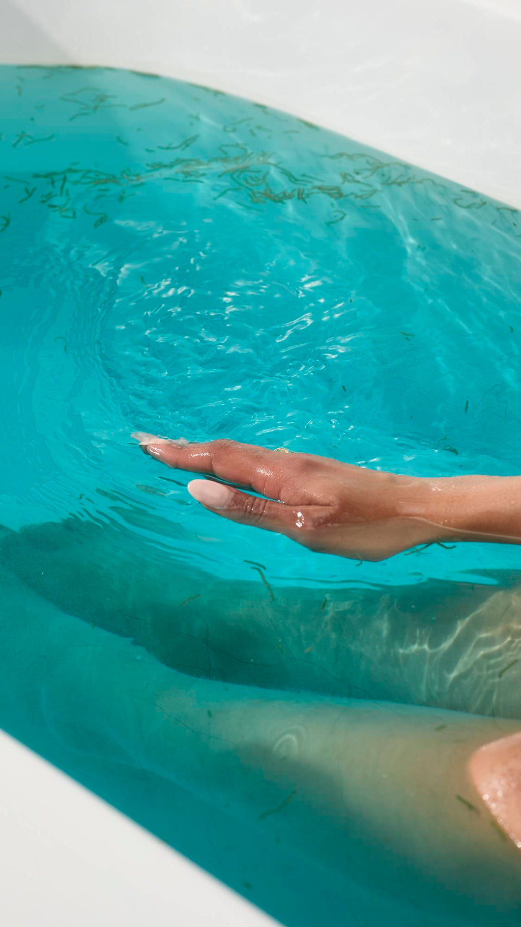 A hand with pale pink nails skims turquoise water filled with seaweed from the Big blue bath bomb. Submerged legs are visible in the background of the bright, ocean-like bathwater.