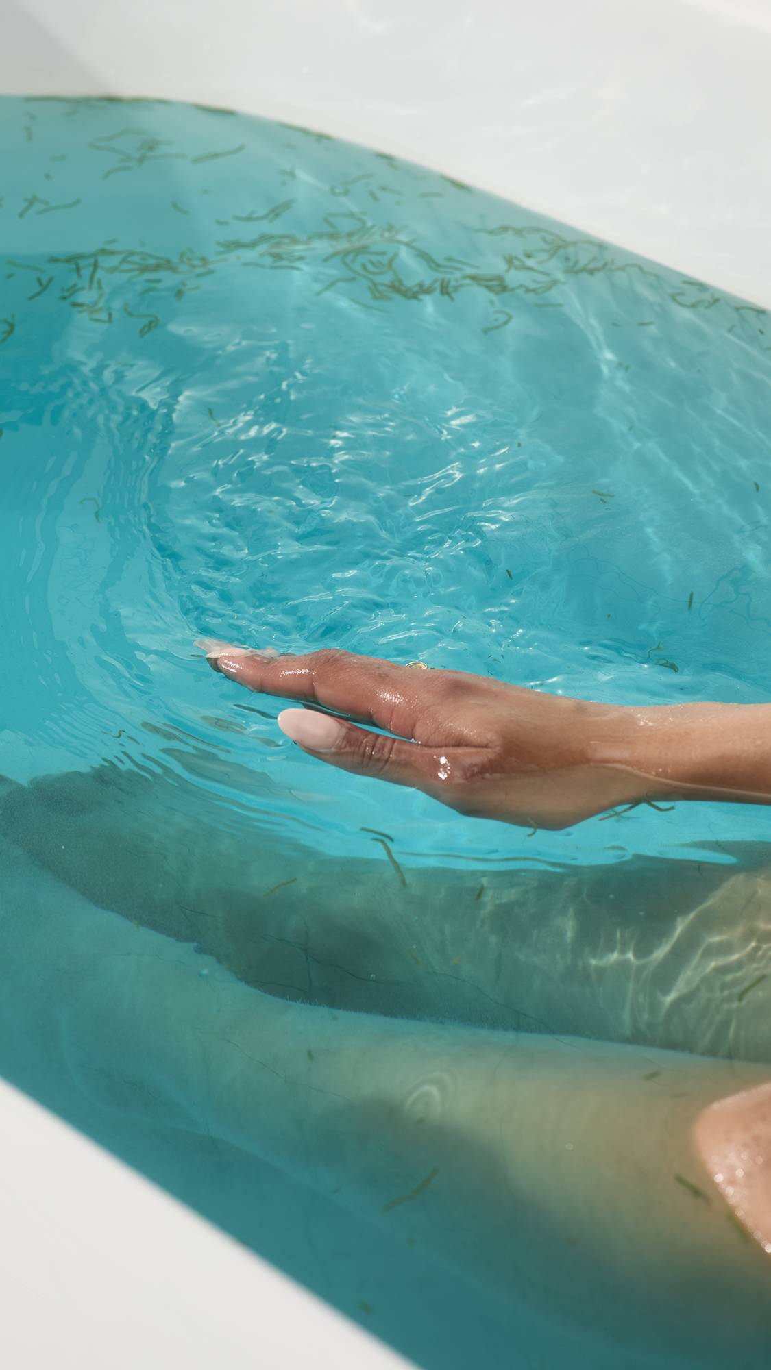A hand with pale pink nails skims turquoise water filled with seaweed from the Big blue bath bomb. Submerged legs are visible in the background of the bright, ocean-like bathwater.