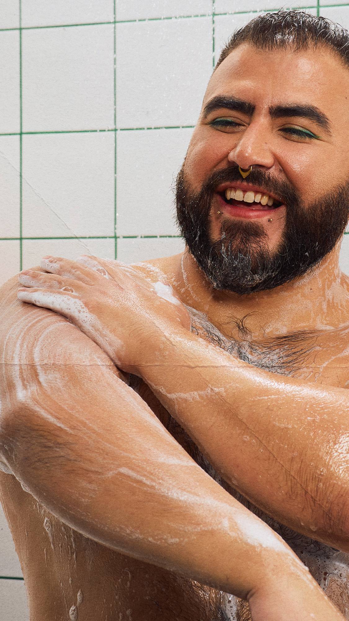 A smiling, bearded model with green eyeliner washes their arm in the shower. They rub thick, white "BOO!" shower gel lather onto their wet skin. White square wall tiles with green grout form the background.