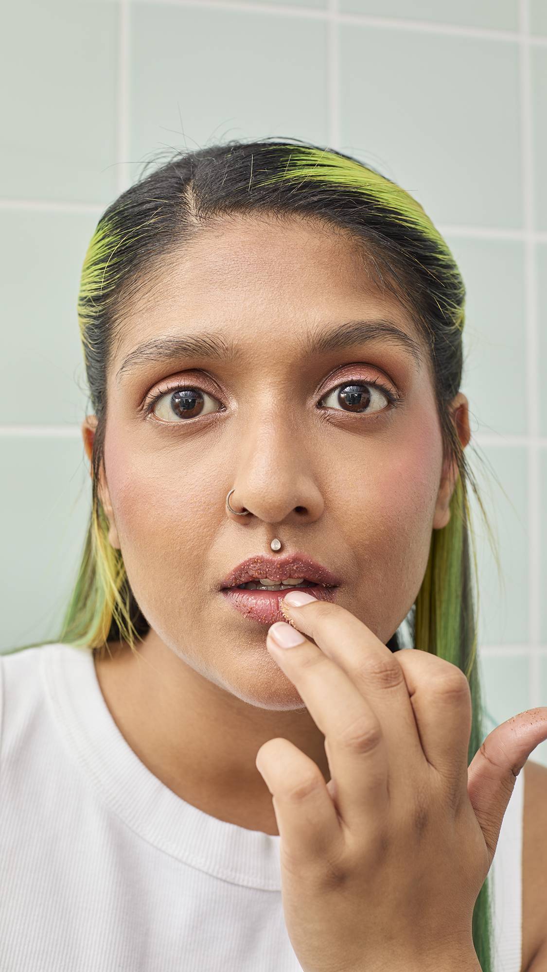 A close-up, front-on portrait of a model with long, bright green hair applying Lush "Buzz" Lip Scrub. They use a finger to rub the brown, sugary product onto their lips while looking directly into the camera against a green-tiled background.