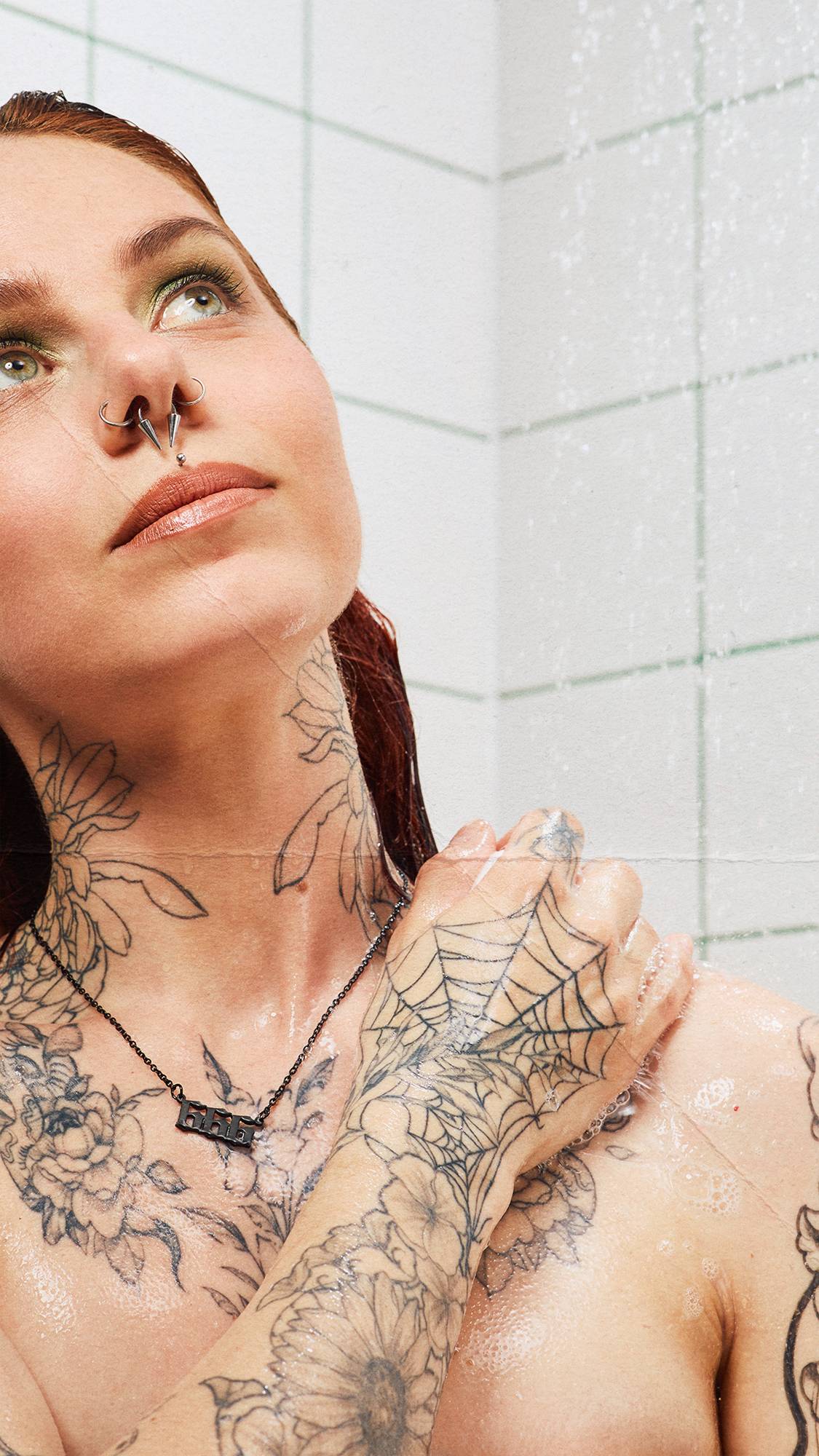 A close-up shot of a tattooed person washing their shoulder under streaming shower water, looking upwards. White wall tiles with green grout form the background.