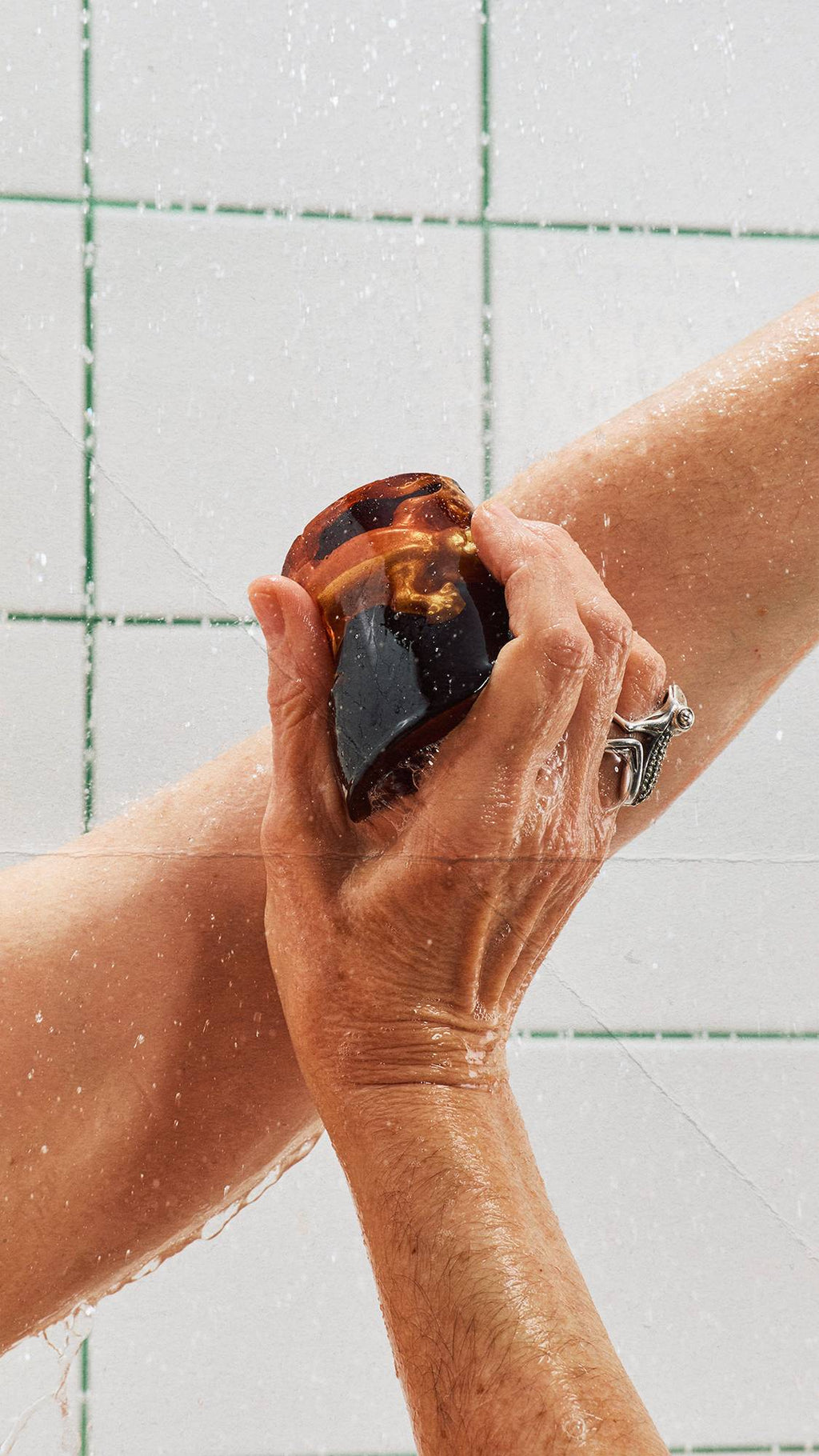 A person washes their arm with a dark, translucent shower jelly containing an embedded golden lizard gummy under streaming shower water. White square wall tiles with green grout form the background.