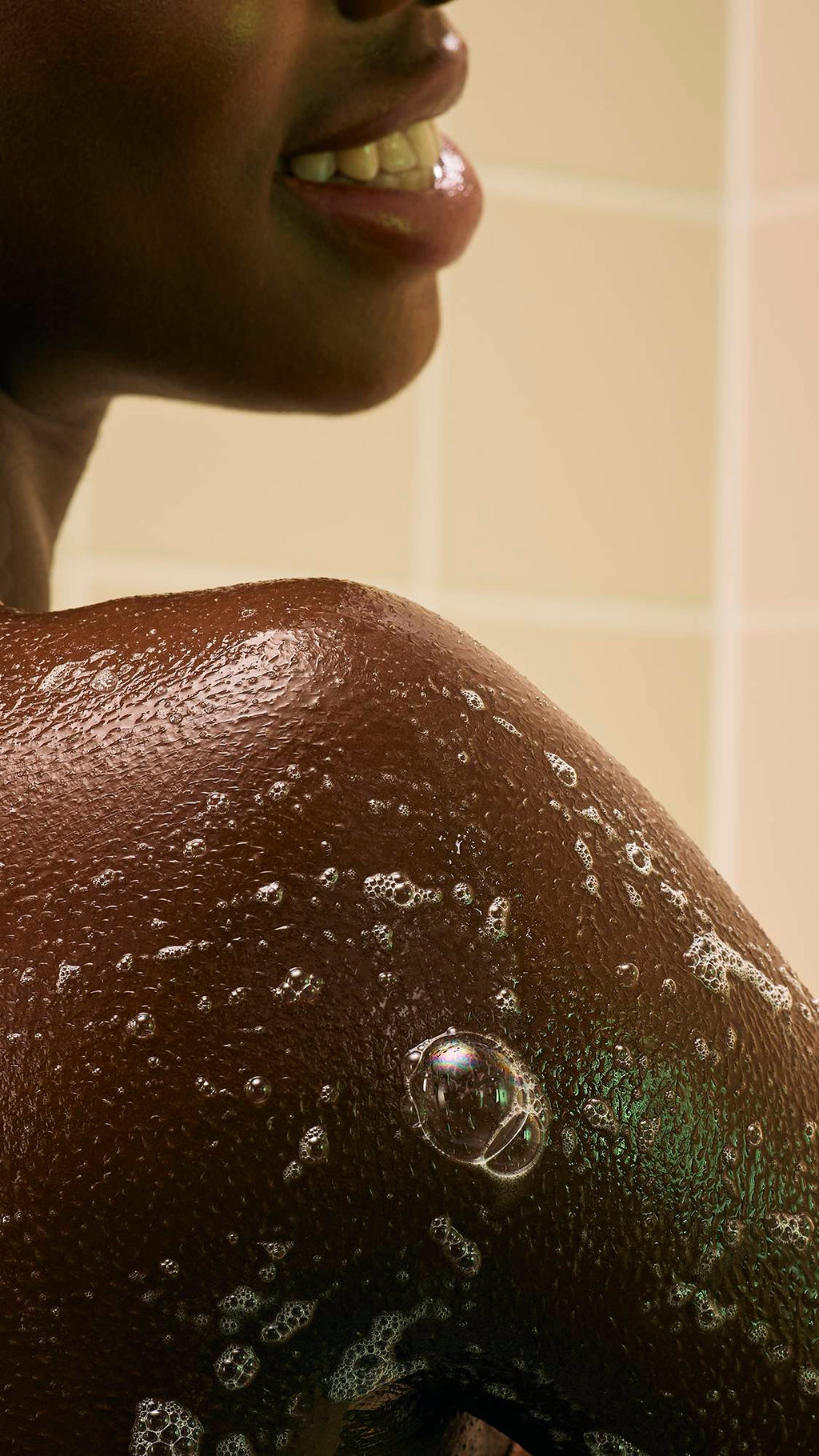 A close-up of wet, glistening skin covered in fine white soap bubbles created by "The Good Hour" shower jelly product. The profile of a person's chin and a smiling mouth are visible at the top,